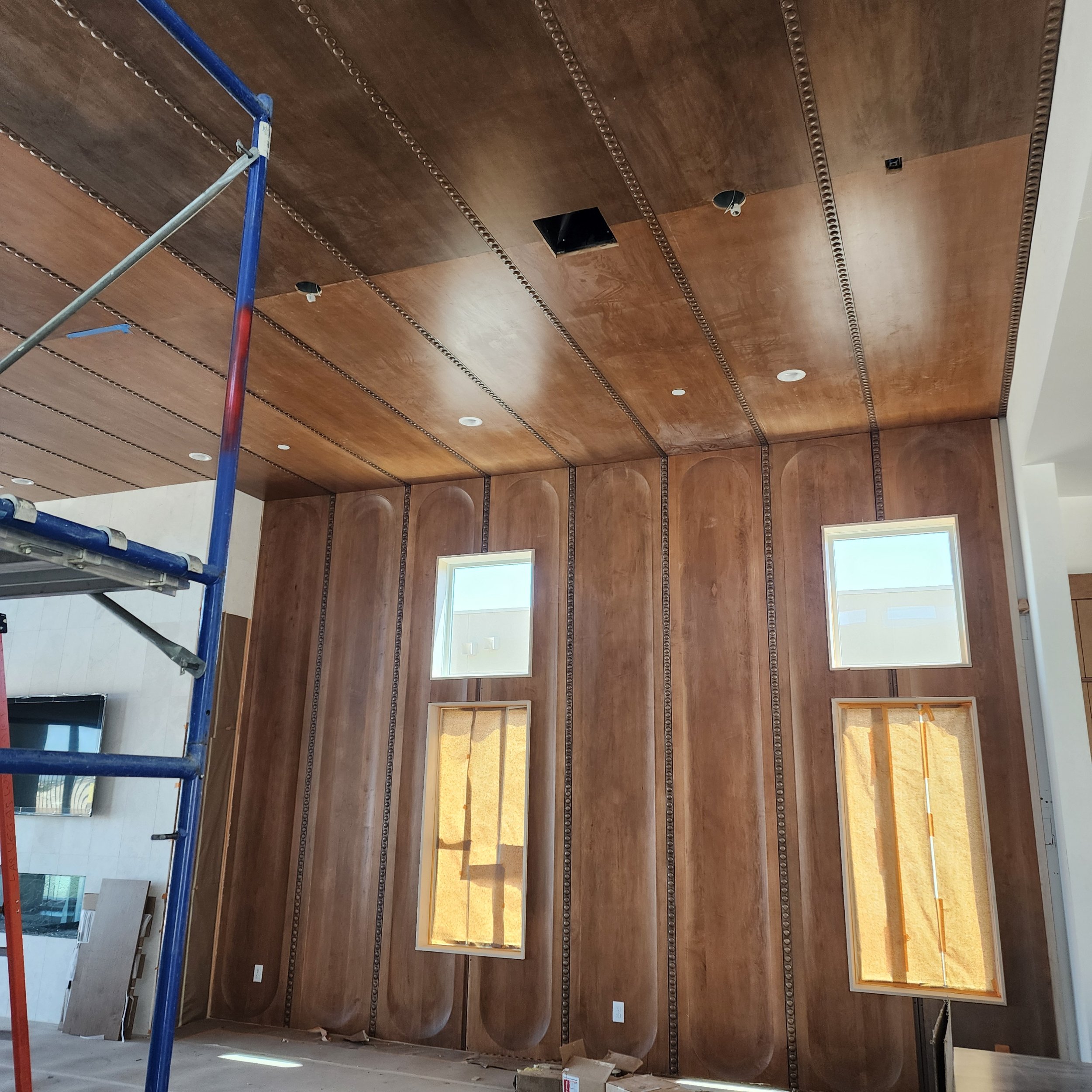 An interior room under construction with a partially installed wooden ceiling featuring decorative molding, with two rectangular windows and a construction scaffold visible.