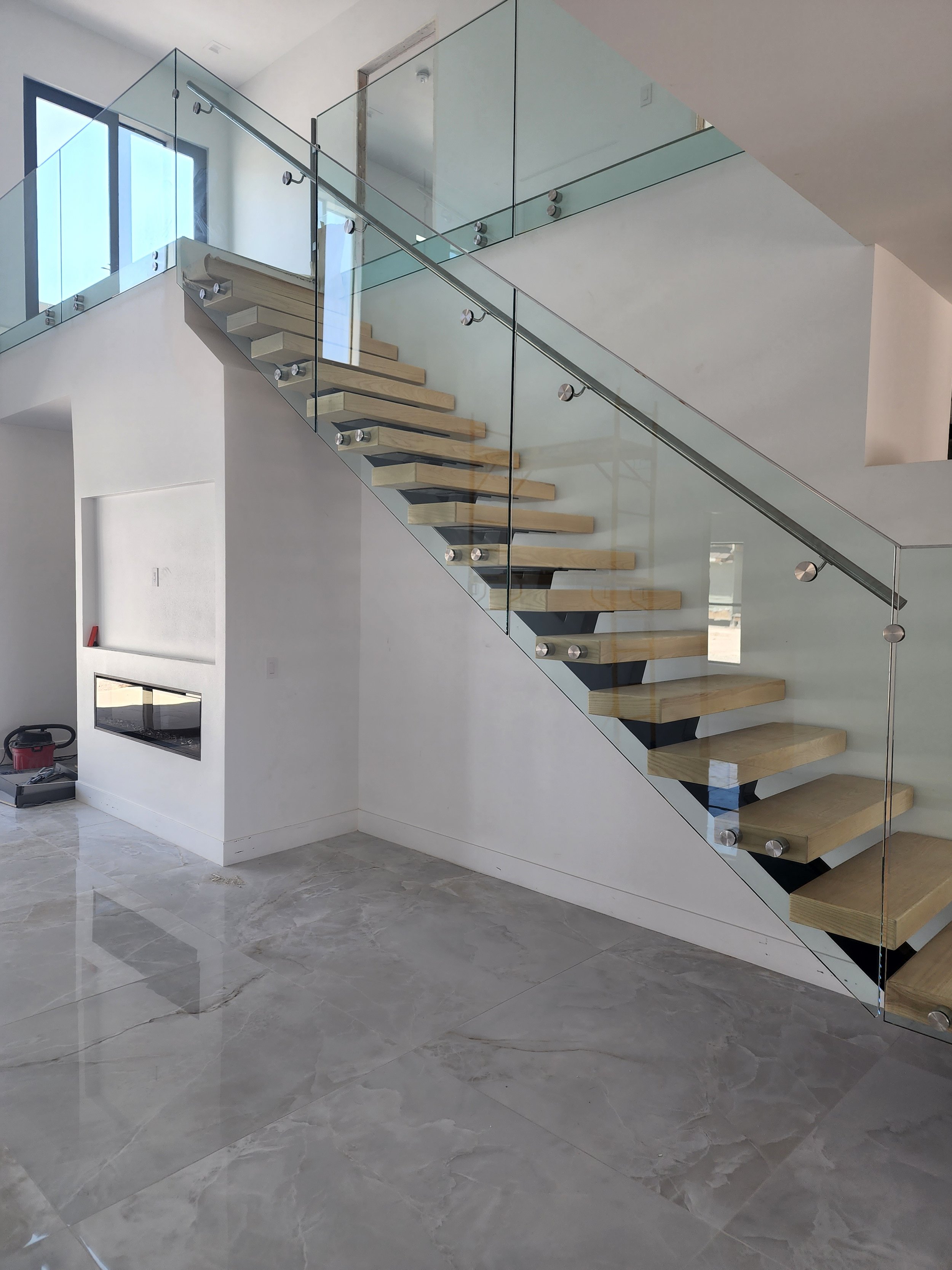 Modern staircase with wooden steps, glass railing, and metal supports inside a bright, minimalistic home with white walls and gray marble floors.
