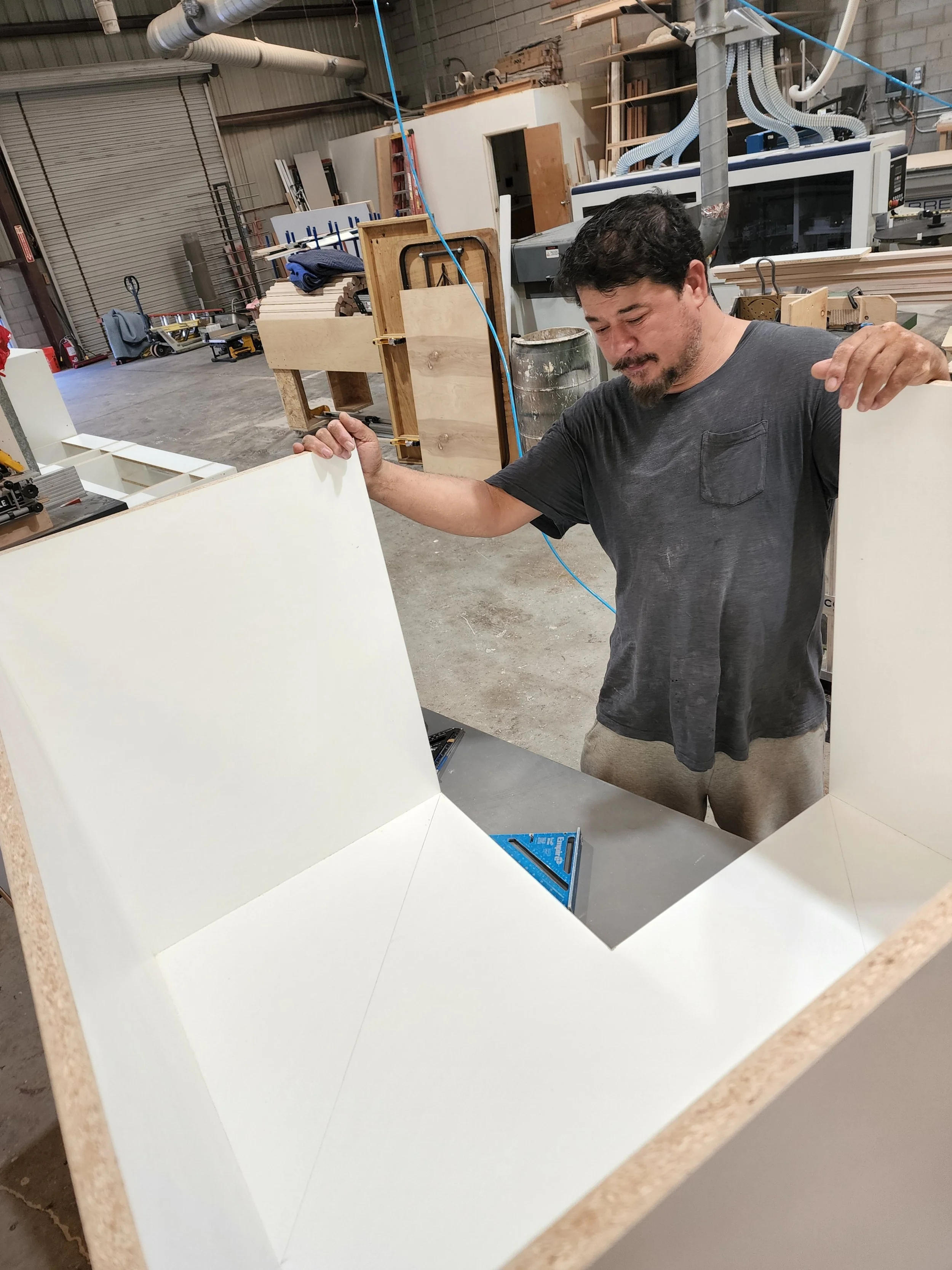 A man working on assembling a white, angular piece of furniture or structure in a workshop with wood and machinery.