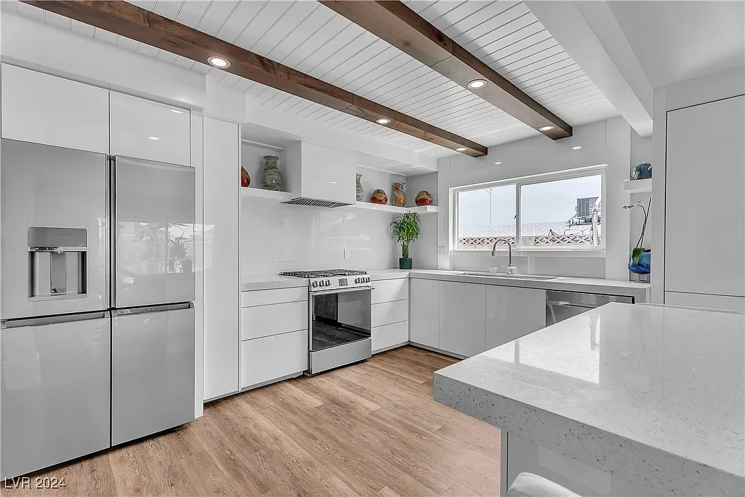 Modern white kitchen with natural wood accents, including stainless steel refrigerator and stove, open shelving with decorative items, a window above the sink, and a white countertop island.