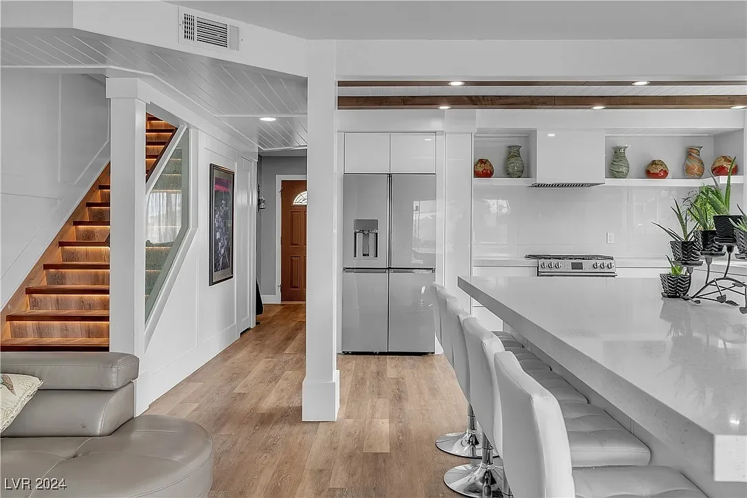 Modern kitchen with white cabinets, a large kitchen island with bar stools, a stainless steel refrigerator, and wooden accents on the ceiling. There are decorative vases on shelves above the stovetop. The space has light hardwood floors and a stairca