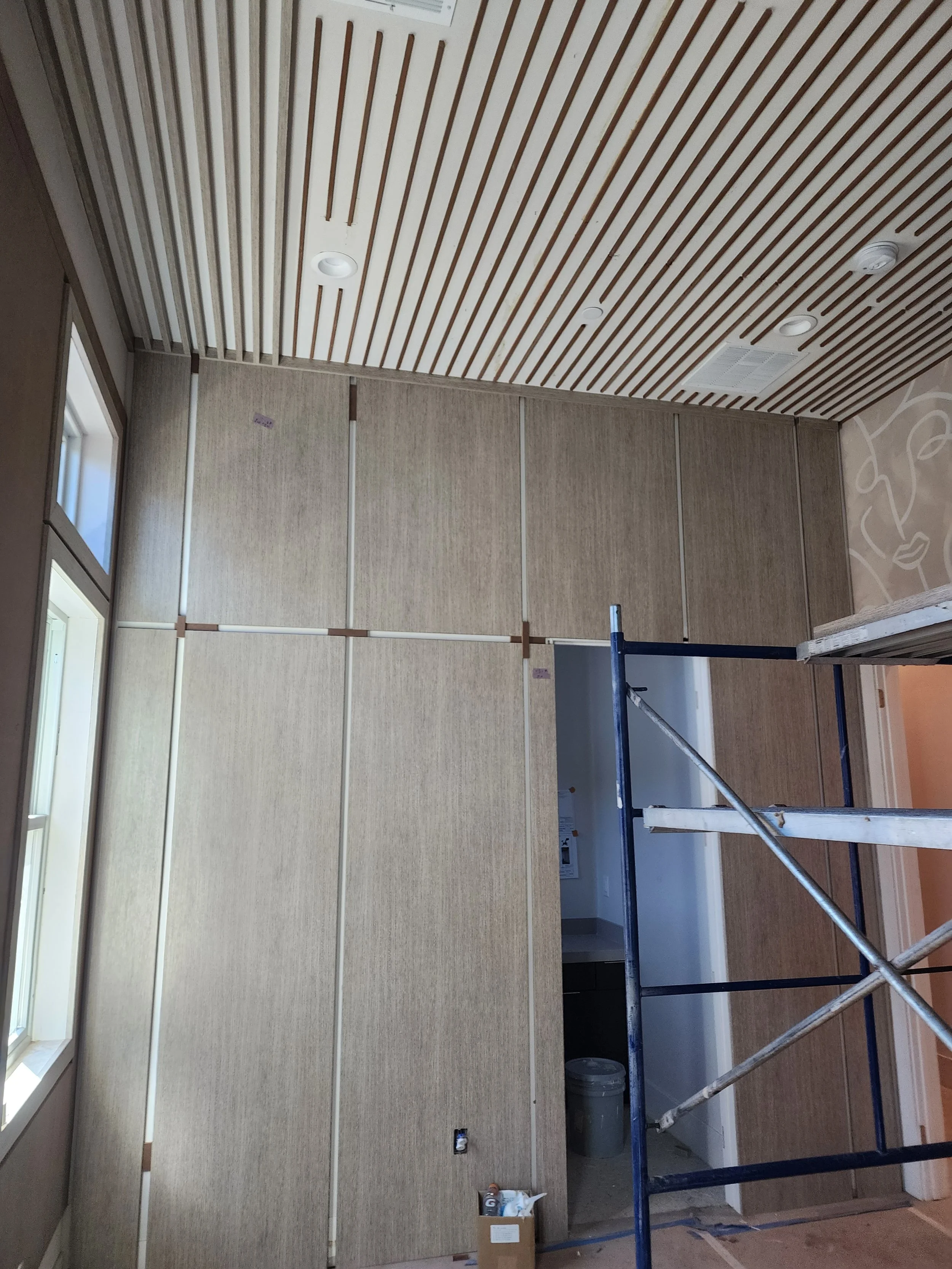 A room under renovation with a partially installed wall covering, scaffolding for workers, a box of supplies, a trash bin, and a window on the left side.