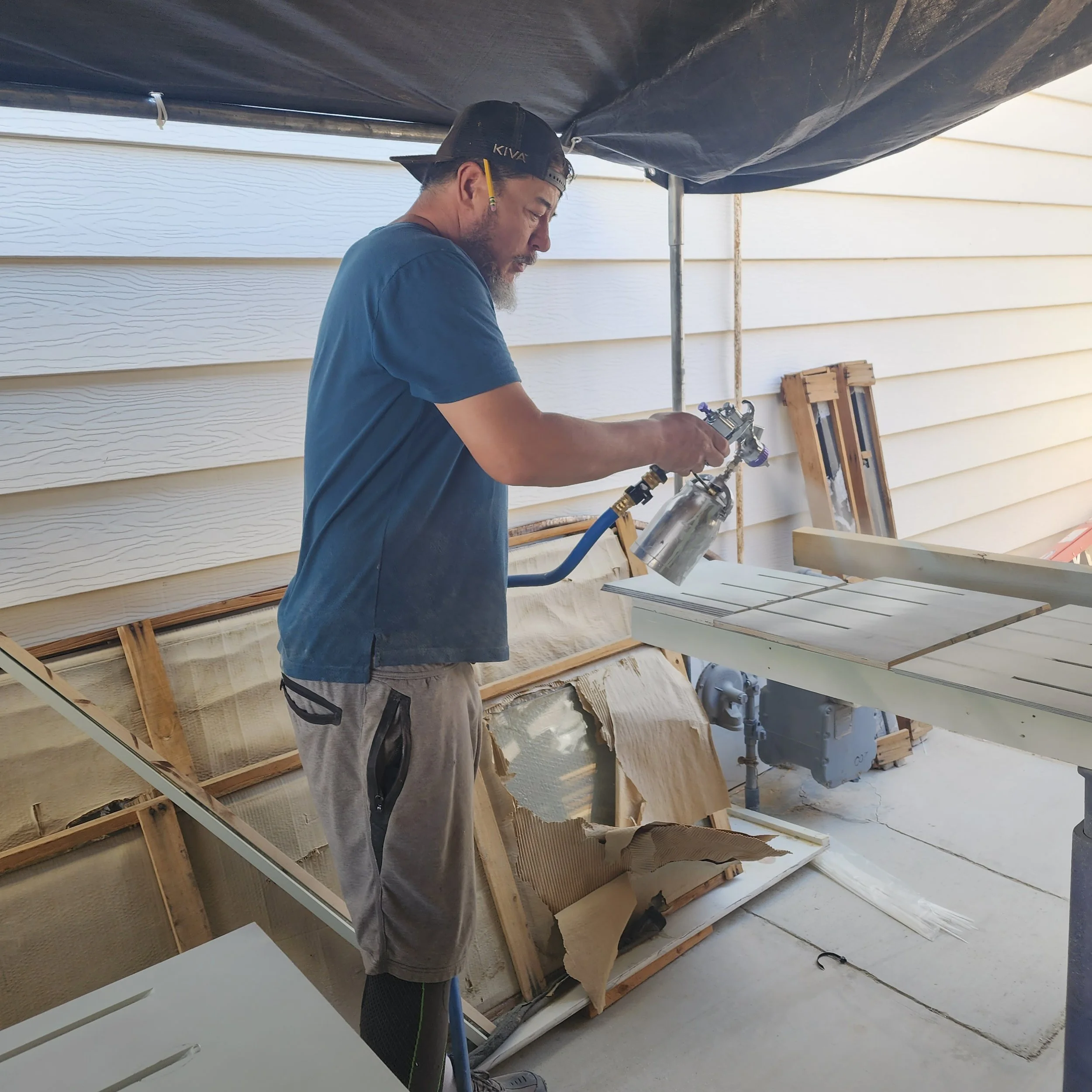 A man wearing a black backward baseball cap, a blue t-shirt, and gray pants is working on a construction project outside. He appears focused as he handles tools or equipment near a wooden structure against a white house siding background. The scene i