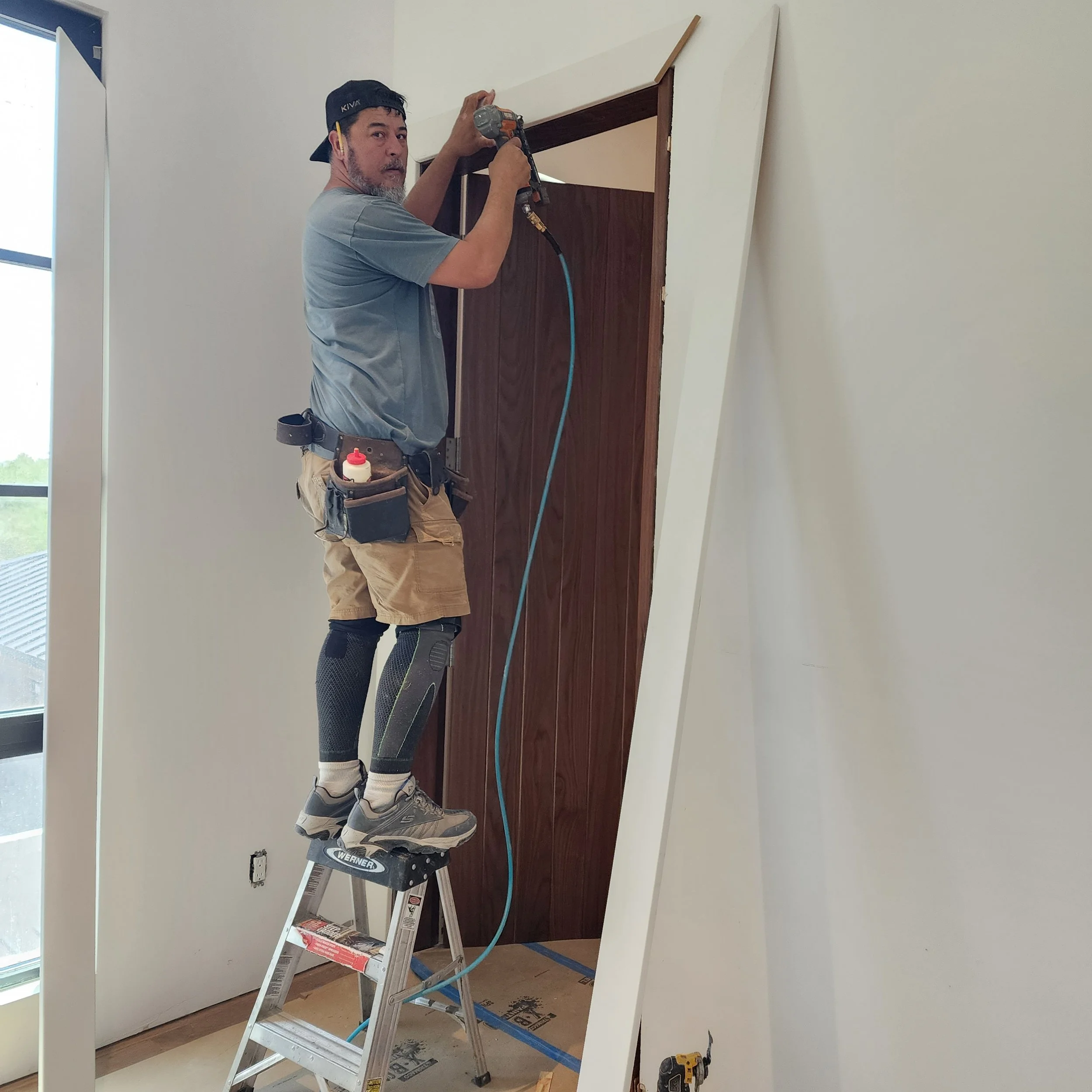 A man standing on a ladder installing or repairing a door or wall panel in a room with renovation materials around.