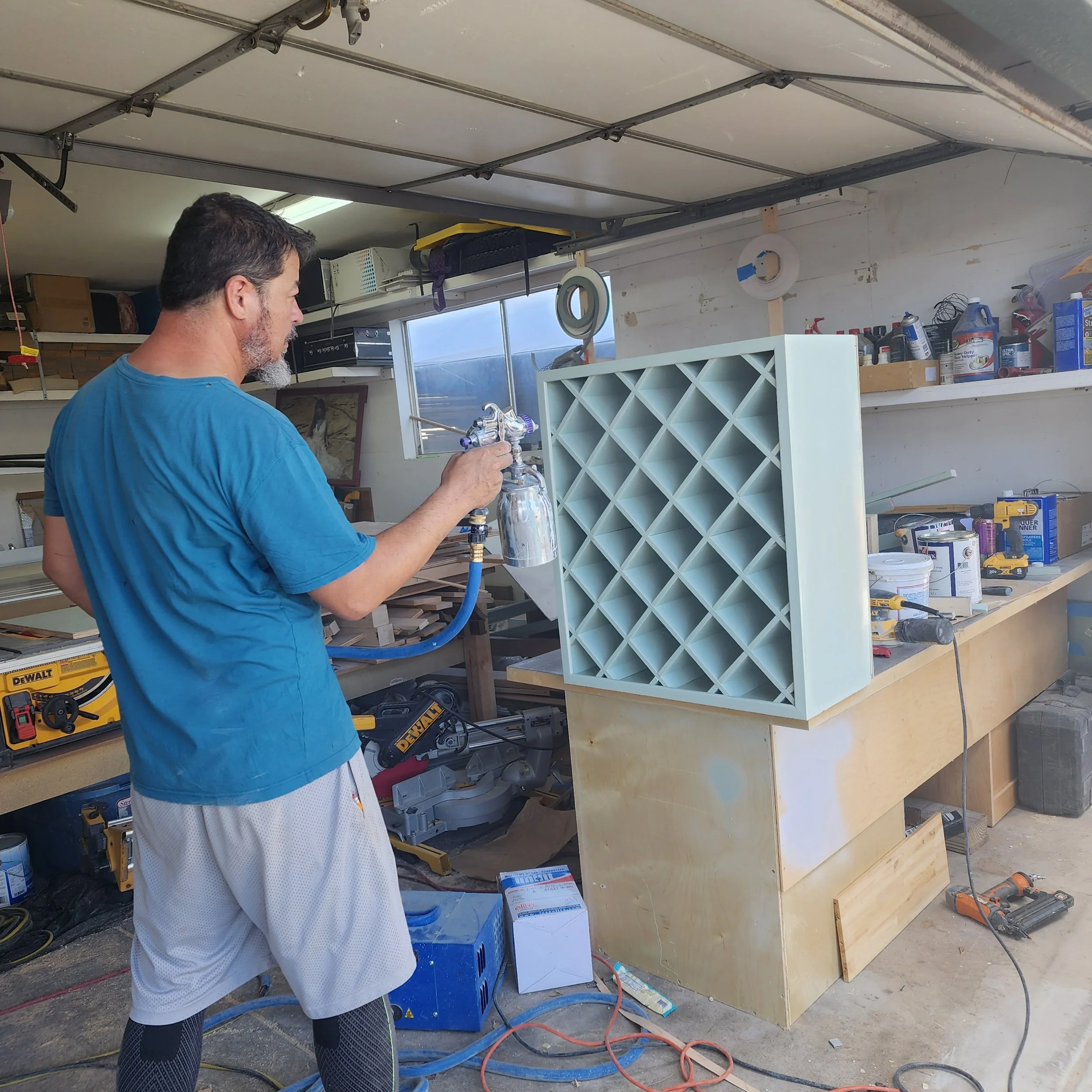 A man working in a garage or workshop, holding a spray gun aimed at a clear plastic or glass object, with various tools and supplies on shelves behind him.