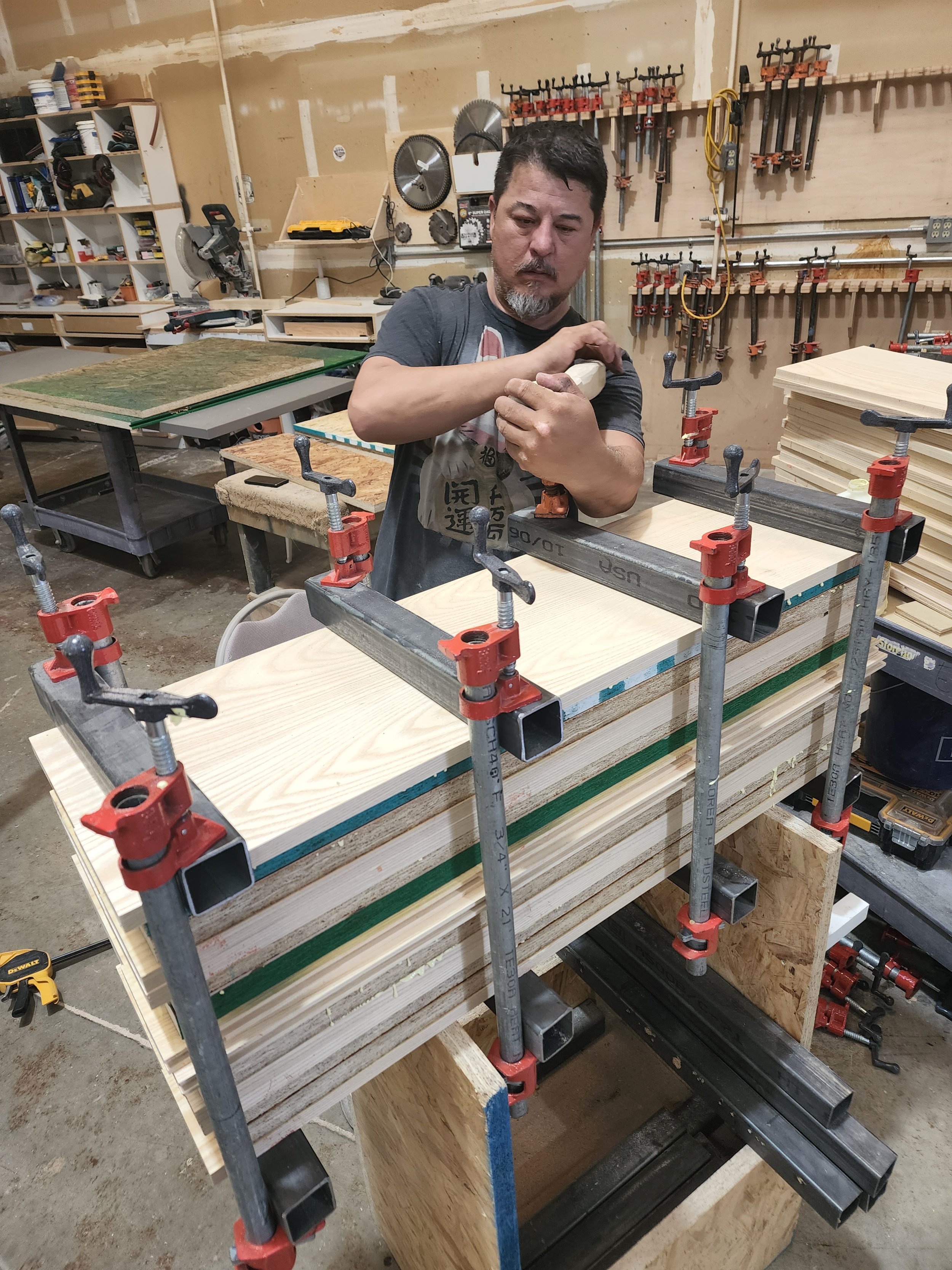 A man in a workshop or garage working on a woodworking project, using clamps to secure a piece of wood on a workbench. The background features various tools on the wall and shelves filled with supplies and equipment.