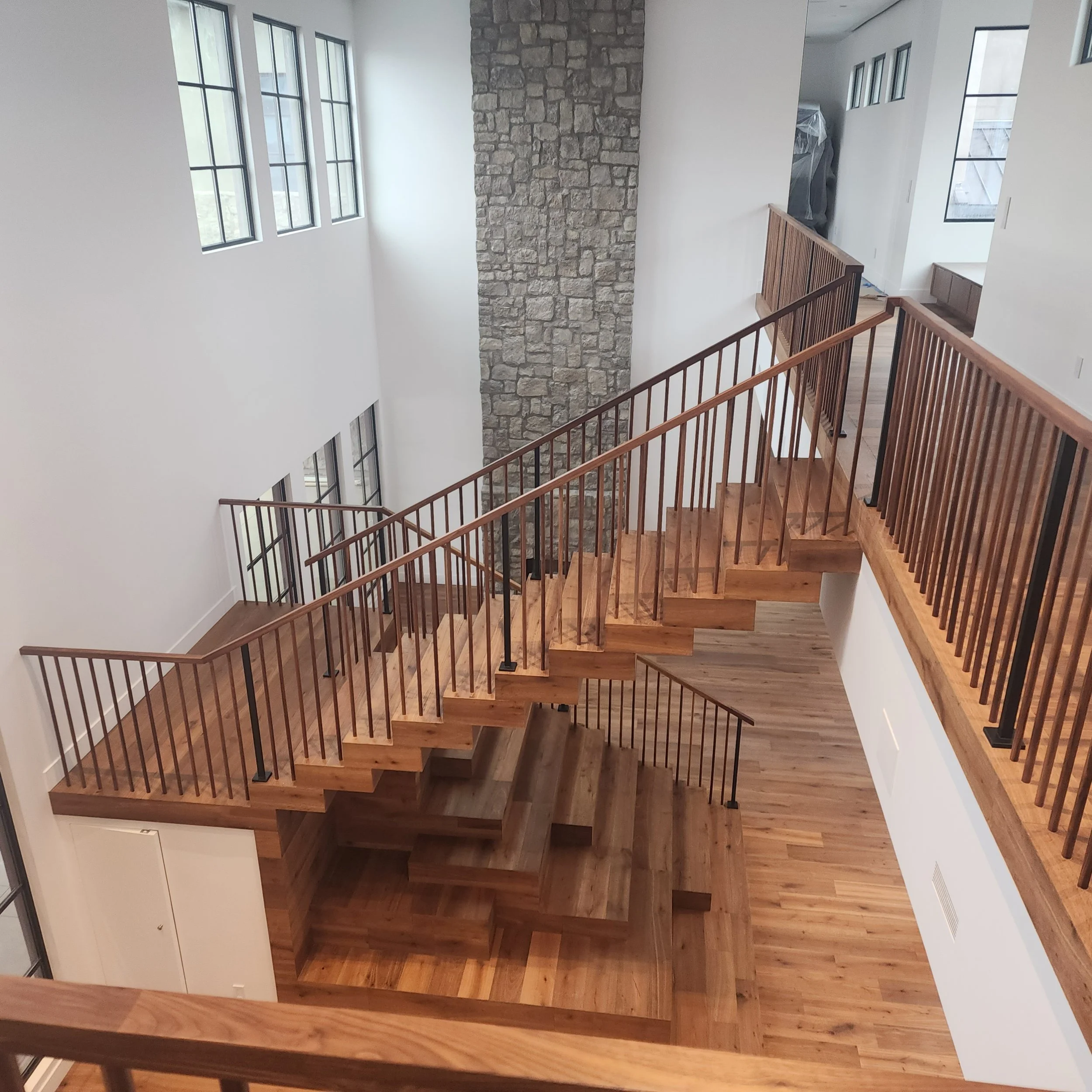 Interior view of a modern house staircase with wooden handrails and flooring, large windows allowing natural light, a stone accent wall, and white walls.