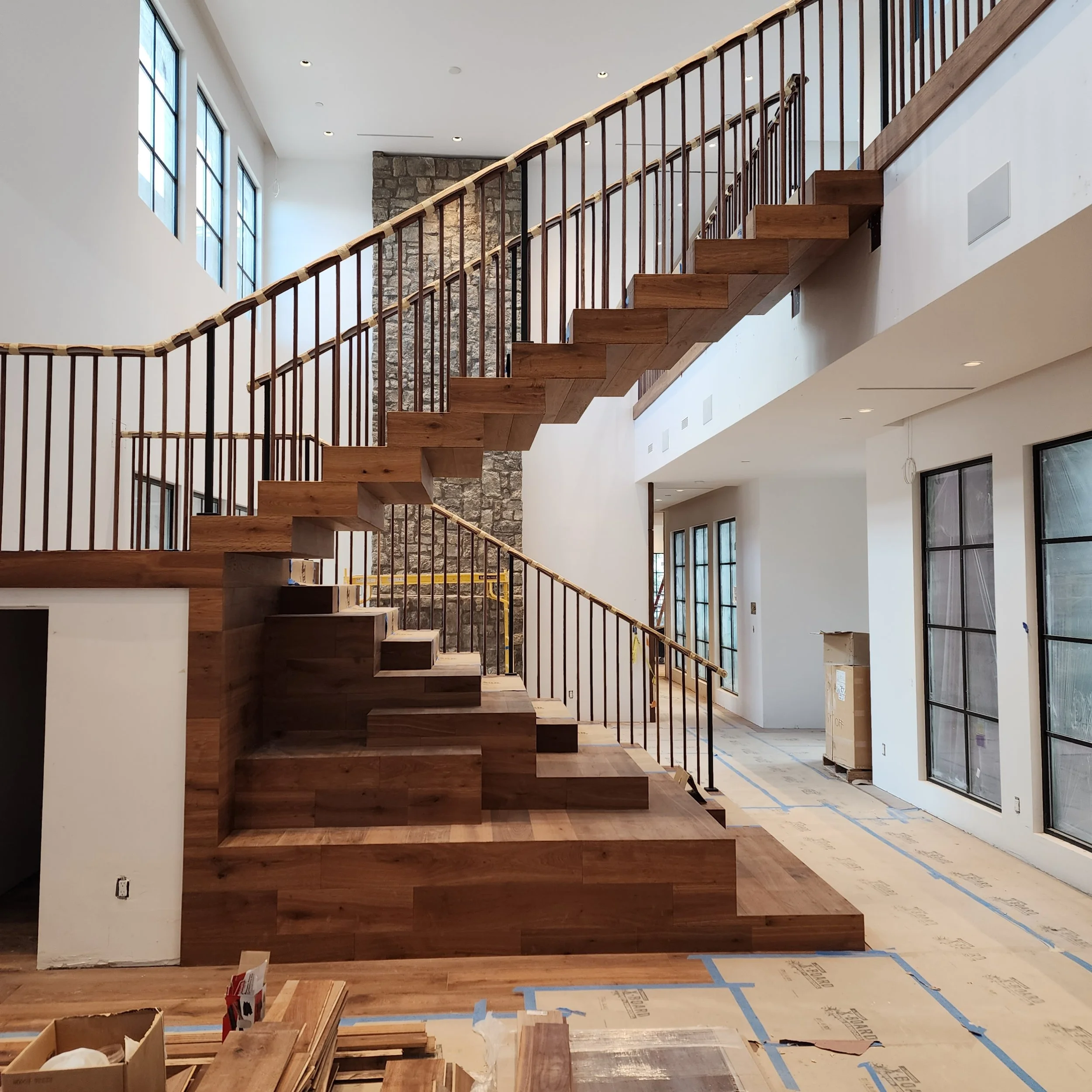 Interior view of a modern house under construction, featuring a wooden staircase with metal railings, large windows, and a stone accent wall.