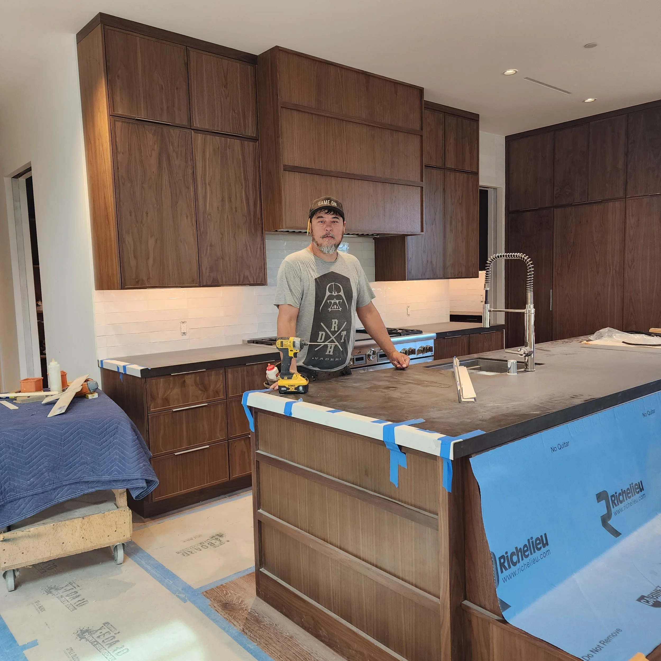 A man standing in a kitchen under construction with dark wooden cabinets and a double sink. He is wearing a gray shirt and a black baseball cap with 'GAME ON' written on it. The kitchen has unfinished flooring and some construction materials.