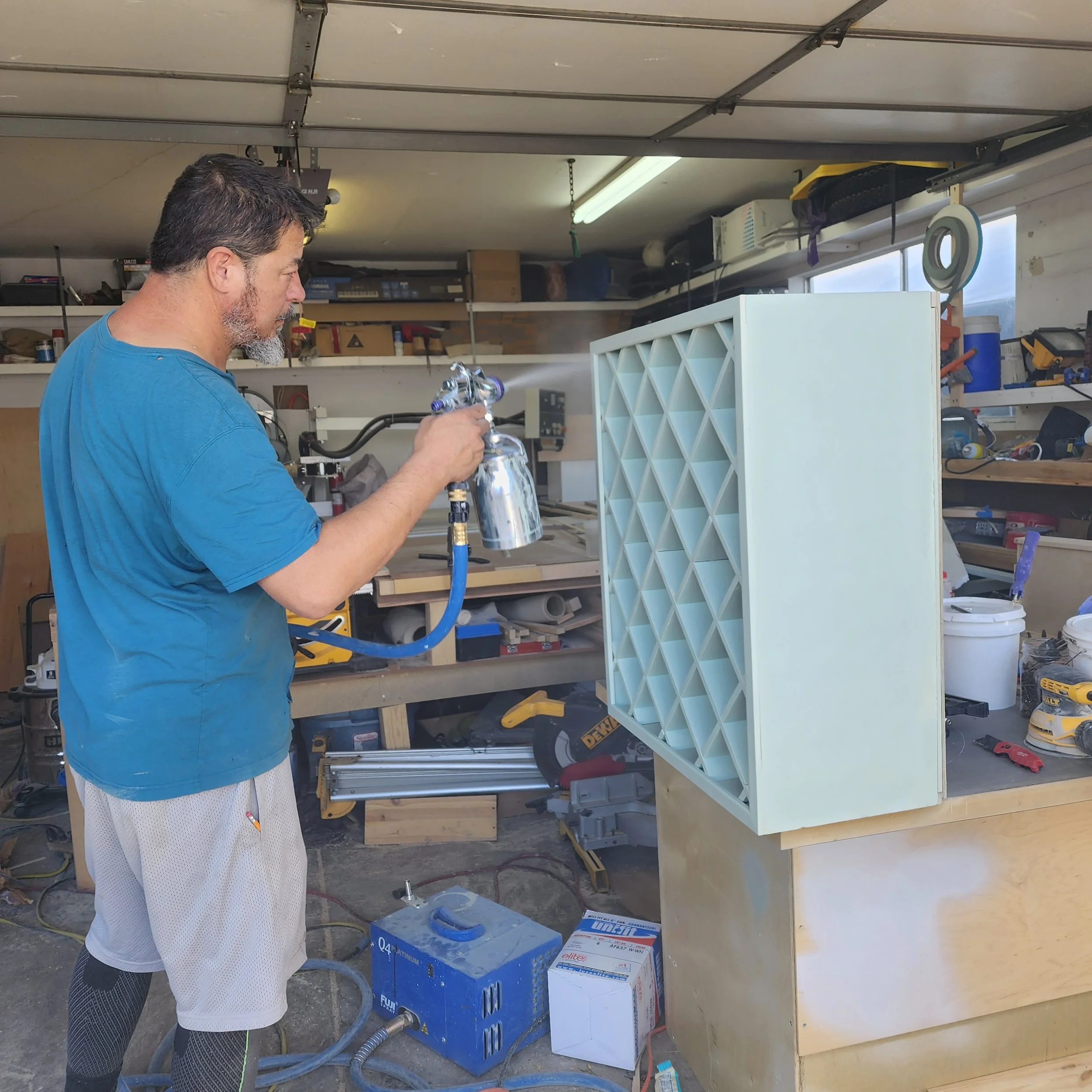 A man in a blue shirt and white shorts in a garage workshop, spray painting a metallic object with a spray gun connected to an air compressor. Shelves with tools and boxes are visible in the background.