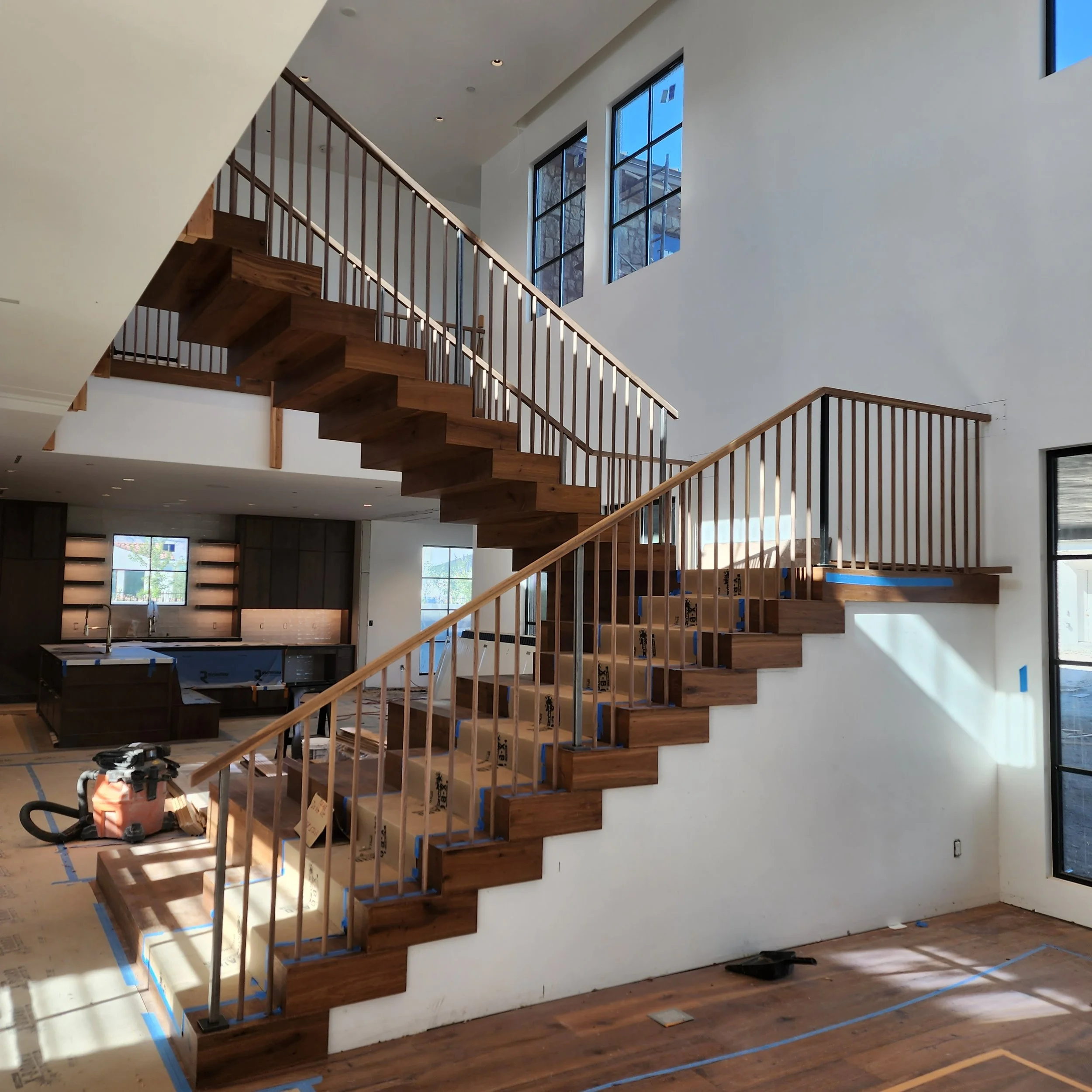 Interior view of a house under construction, showcasing a wooden staircase with black metal railing, large windows, and a dark kitchen area in the background.