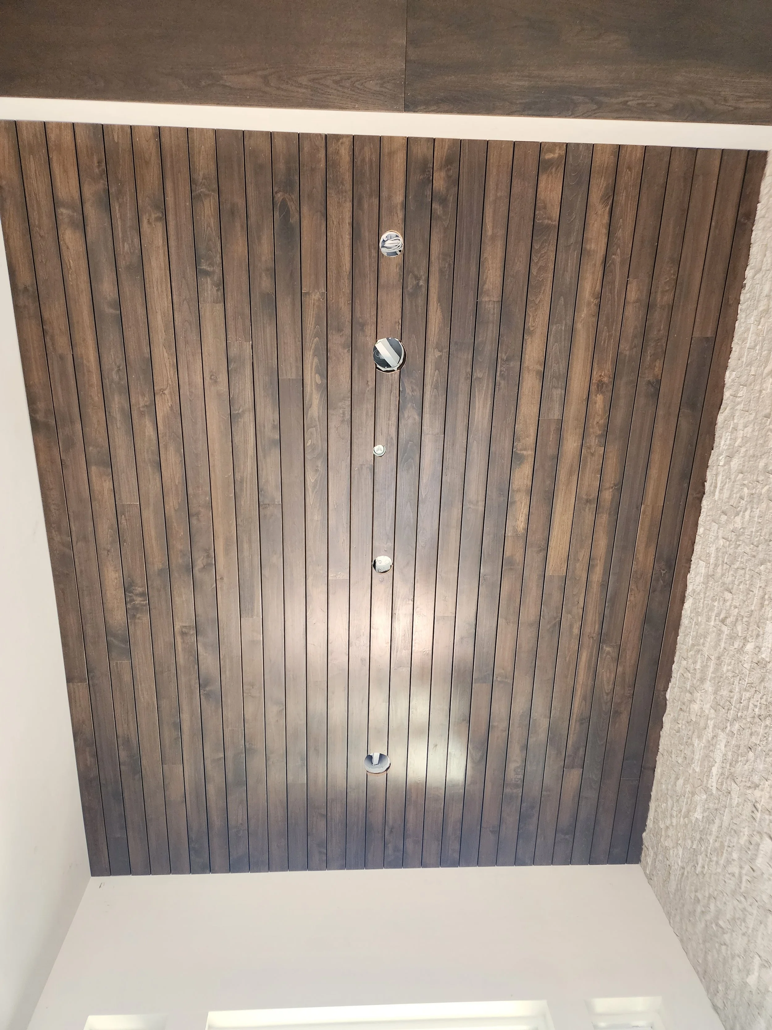 Close-up view of a wooden ceiling with round recessed lights, and part of a light-colored wall and textured carpeted flooring.