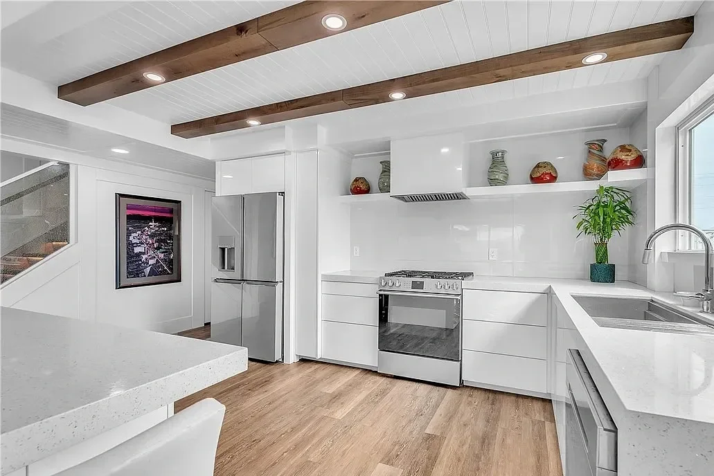 Modern white kitchen with wooden ceiling beams, stainless steel refrigerator, stove, and sink, decorated with colorful vases and green plants.