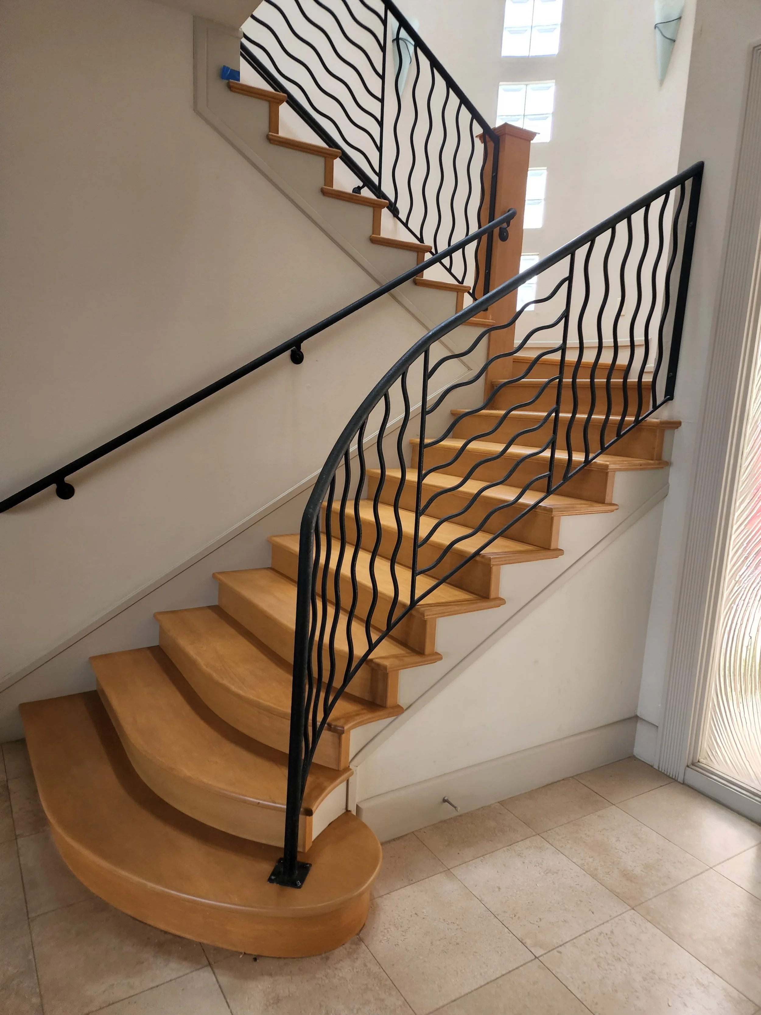A wooden staircase with a curved lower section, light wood steps, black wavy metal railings, and beige walls. There is a window with frosted glass blocks in the background and a mirror on the wall to the right.