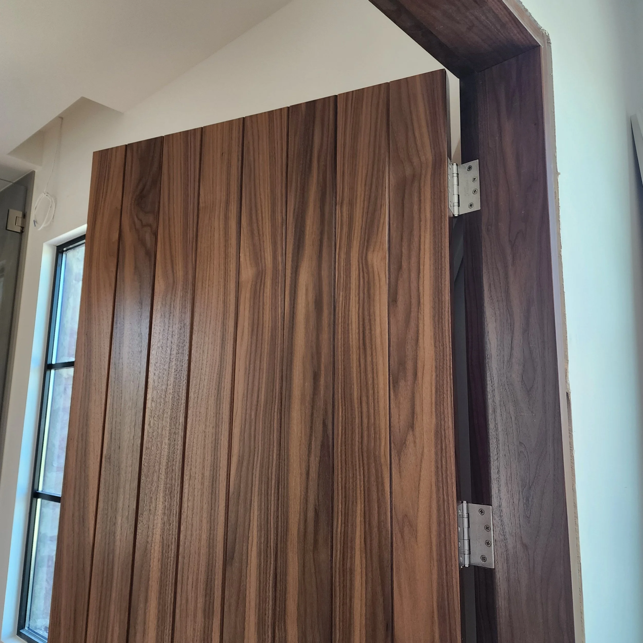The image shows a close-up of a wooden door with visible hinges, set in a wall near a small window with a black frame, and a white wall with a ceiling corner in the background.