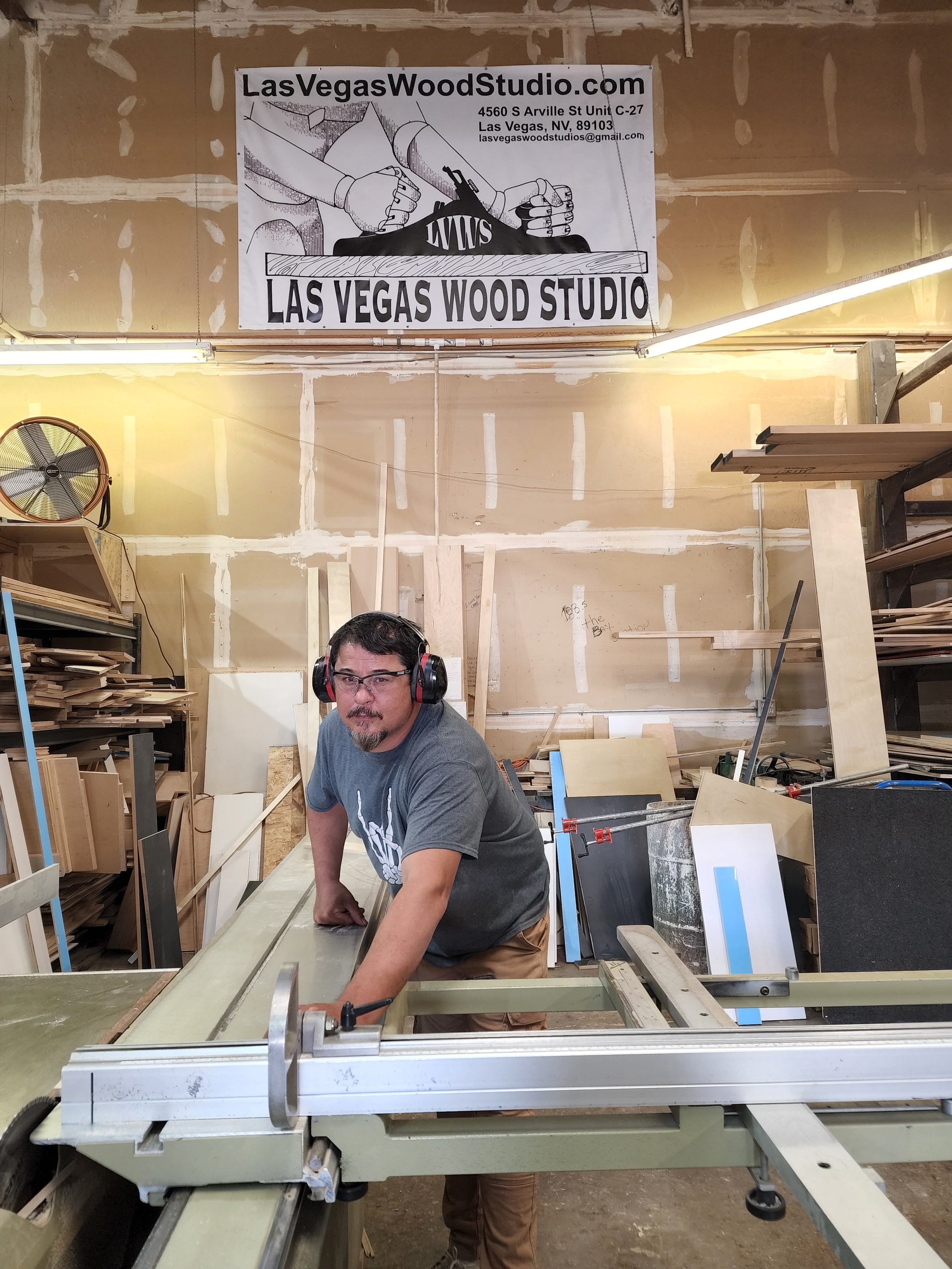 A man wearing safety headphones and glasses working in a woodworking shop. The shop has unfinished walls with a banner that reads 'Las Vegas Wood Studio,' featuring a graphic of hands using a saw and a nail gun. The man is leaning over a large piece 
