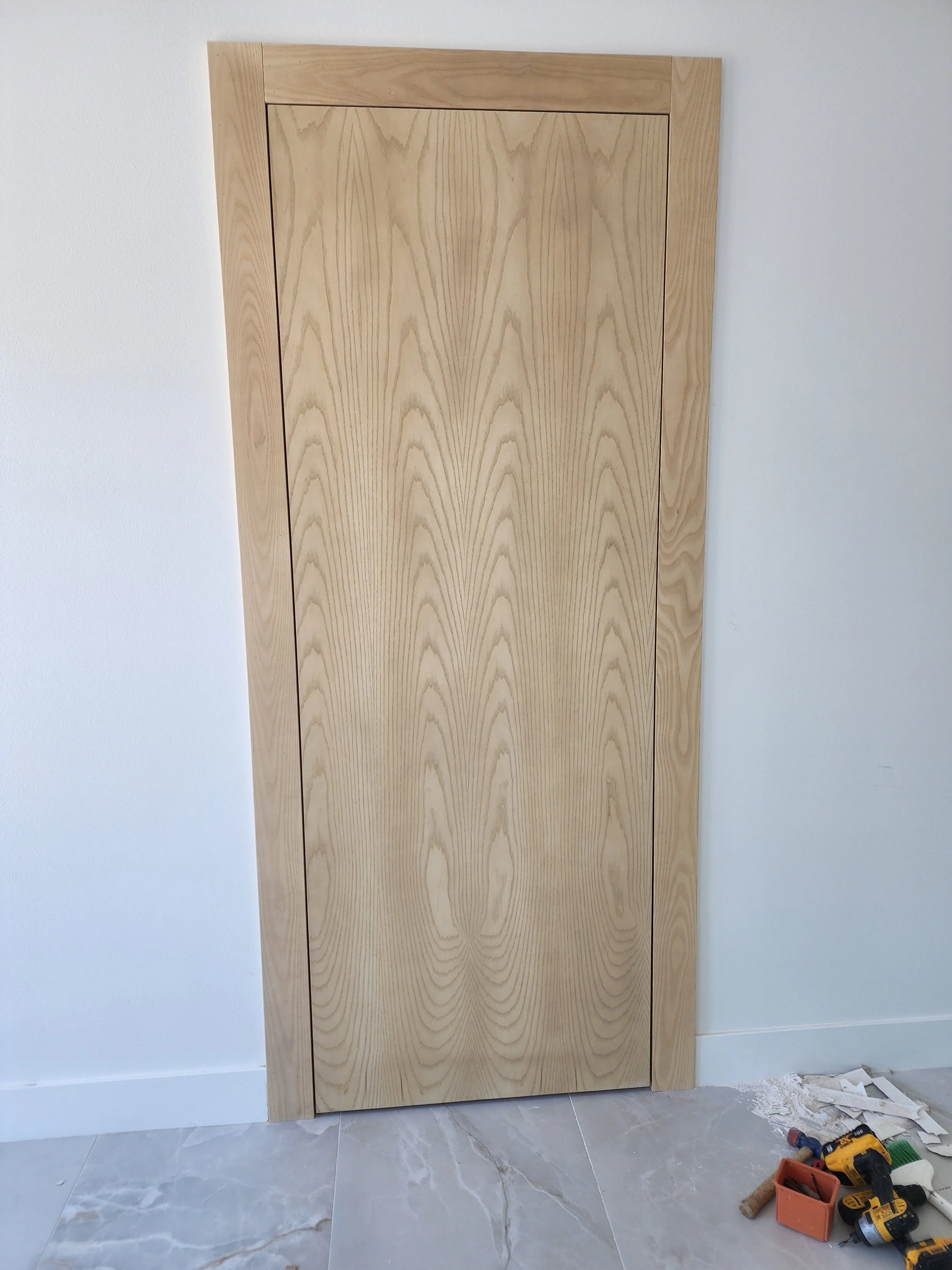 A tall, unfinished wooden cabinet with a natural wood grain finish, against a white wall. Tools and construction supplies are on the floor nearby, indicating ongoing carpentry work.