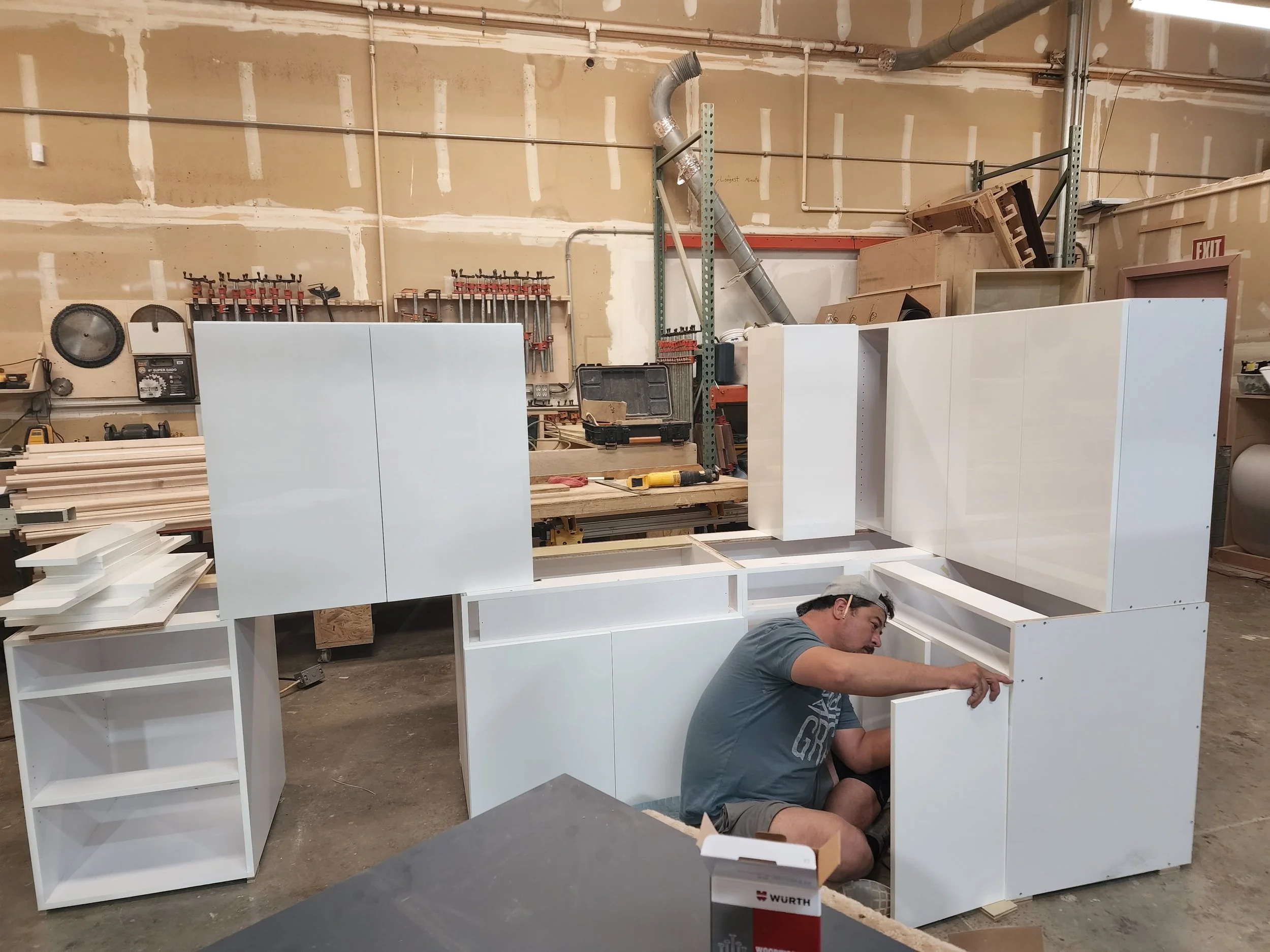 A man working on assembling white wooden furniture in a workshop or garage. The background shows tools hanging on the wall and a partially completed piece of furniture.