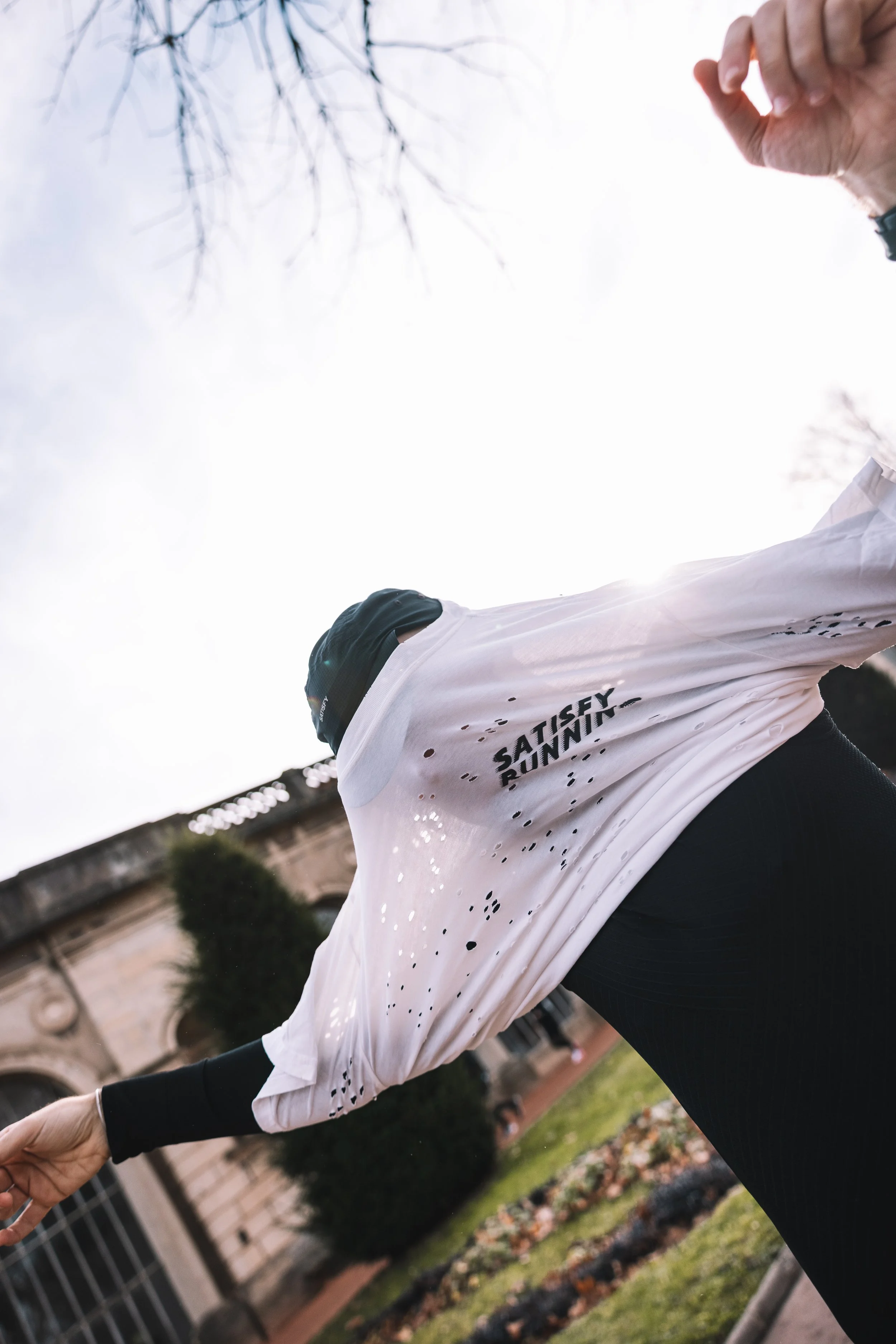 Une personne en tenue de course portant un t-shirt blanc avec le texte "SATISFY RUNNING", faisant un jog dans un parc avec des arbres et un bâtiment en arrière-plan, sous un ciel nuageux. Photo prise au sein du parc de la tete d’or à Lyon. 