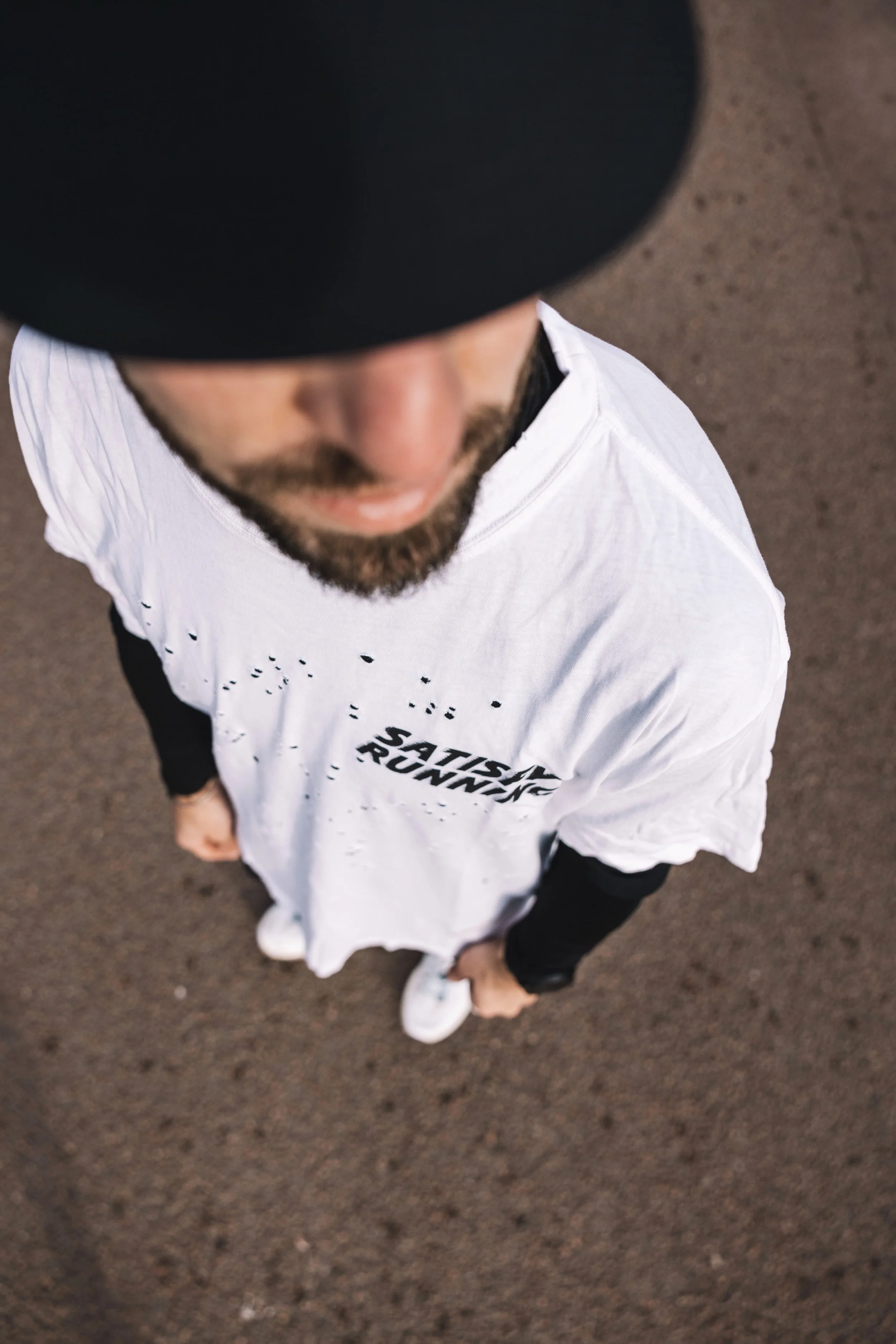 Vue plongeante d'un homme portant une casquette noire, une chemise blanche avec le texte 'Satisfy Running' et des baskets blanches, sur un sol en terre.