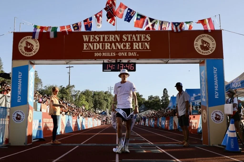 Un homme souriant franchit la ligne d'arrivée d'une course d'endurance de 100 miles, moment capturé lors d'un événement en plein air sous un ciel clair, avec des spectateurs et le logo de la course visible.