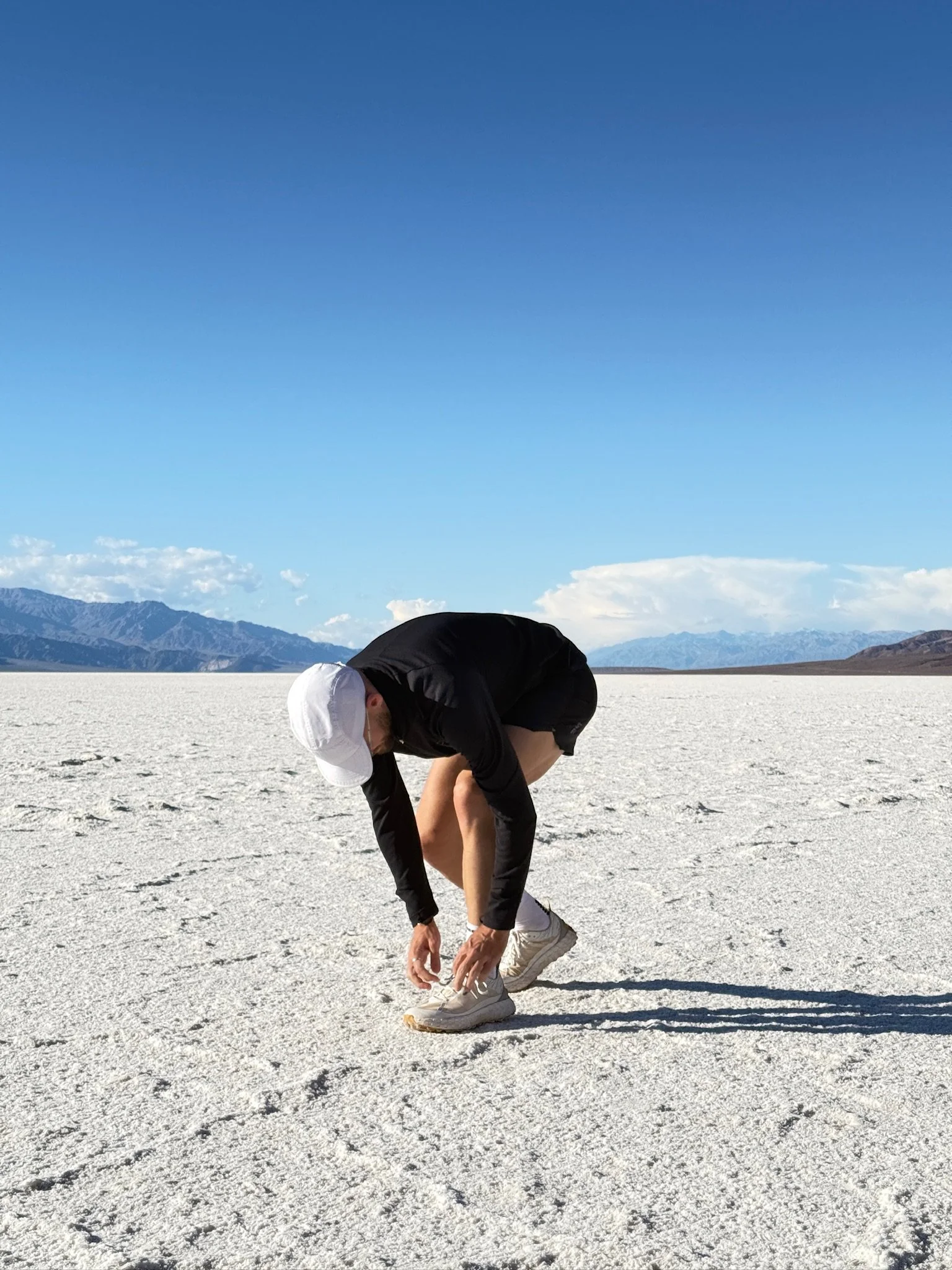 Une personne courbée, en train de attacher ses chaussures dans un paysage désertique avec un sol blanc, des montagnes en arrière-plan et un ciel bleu clair. La photo a été prise à Badwater, dans la Death Valley.