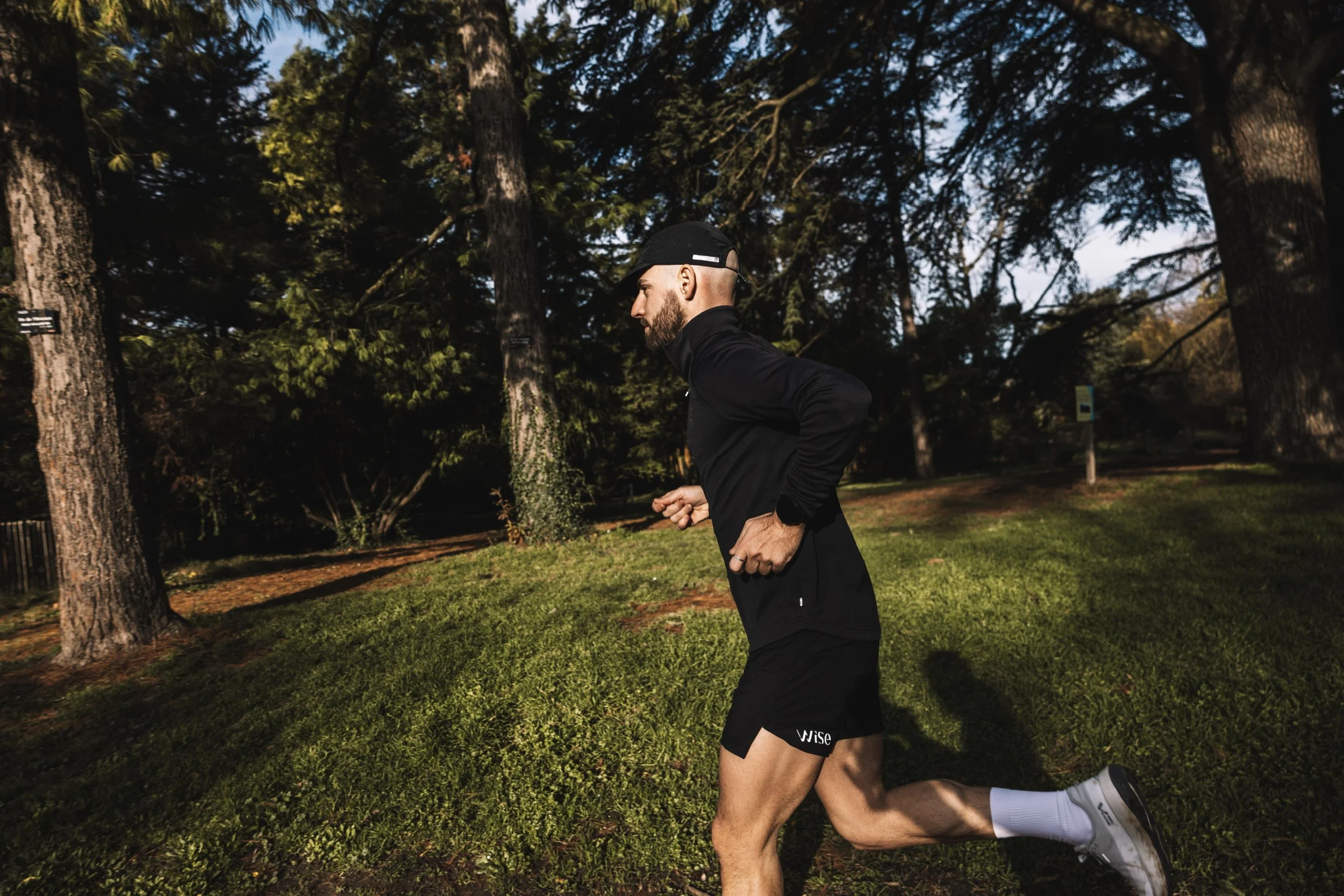 Un homme jogging dans un parc, portant une tenue de sport noire de la marque Wise ultrarunning, un casquette noire satisfy, des chaussures de course blanches Mount to Coast et des chaussettes de running blanches Ravanel.