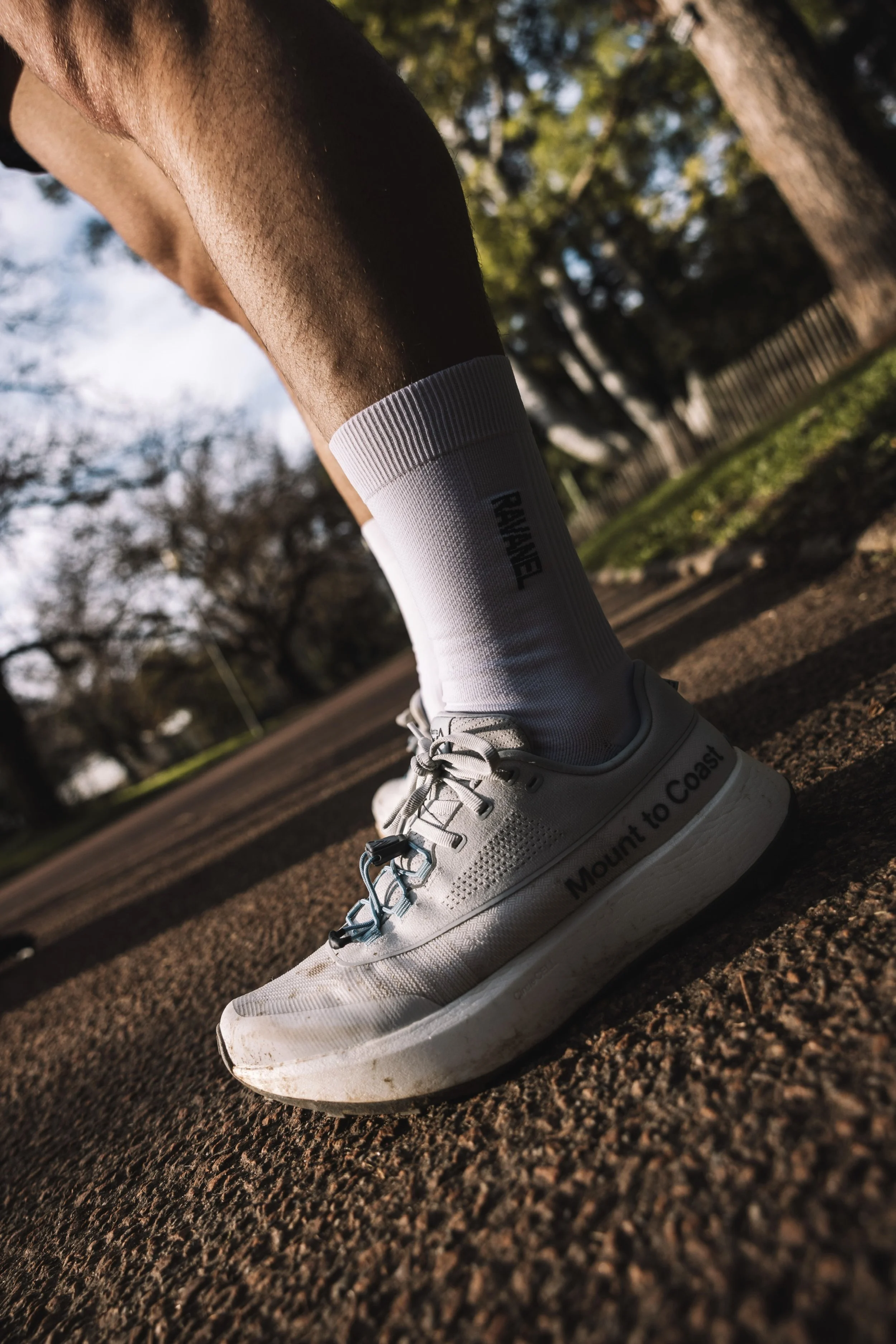 Une chaussure de course blanche avec les mots "Mount to Coast" écrit dessus, portée par une personne portant une chaussette blanche.Surface de course en terre, avec des arbres en arrière-plan sous un ciel ensoleillé.