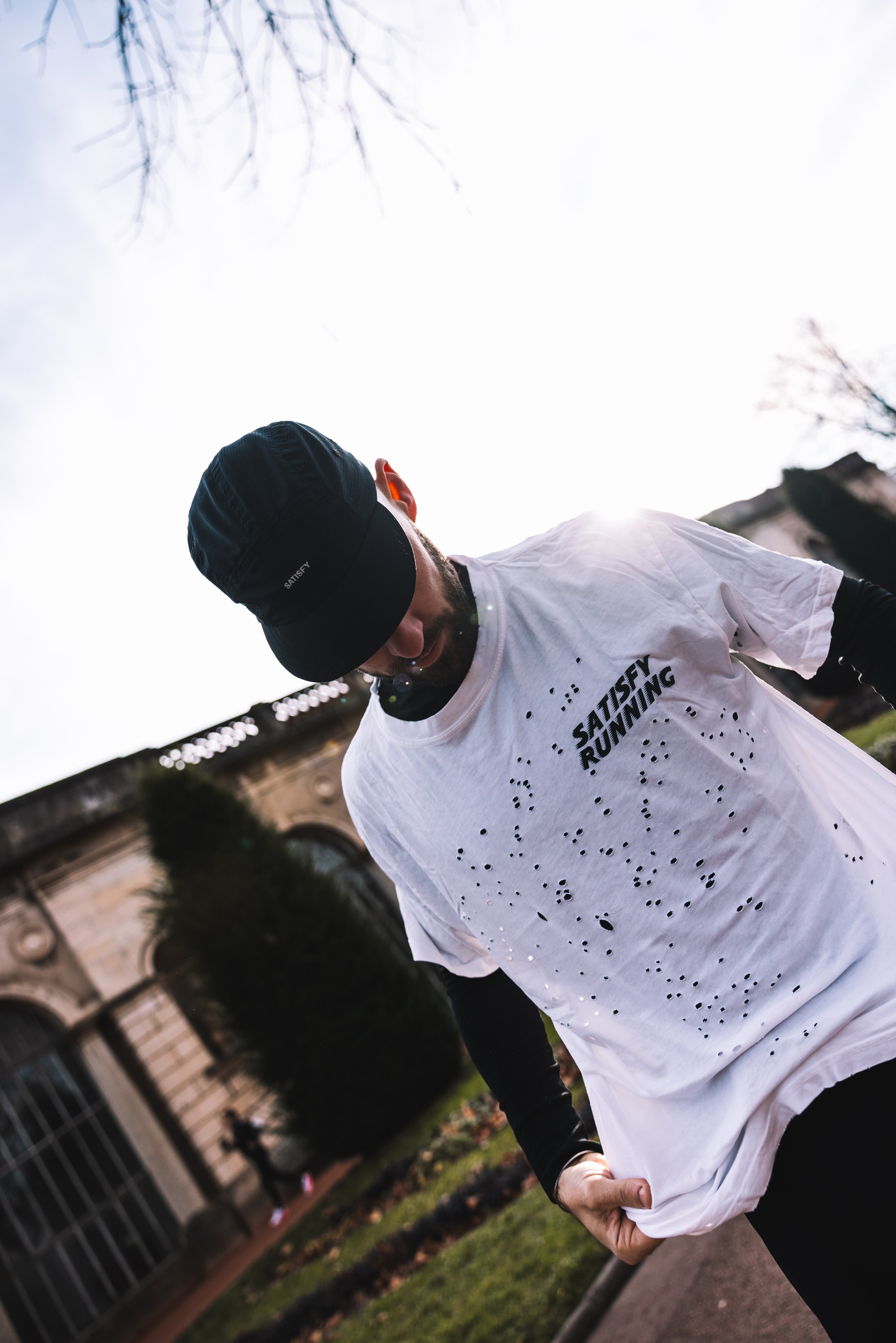 Un homme portant un t-shirt blanc avec des perforations et un béret noir, debout à l'extérieur devant un bâtiment ancien, la tête inclinée vers le bas, dans un environnement urbain.