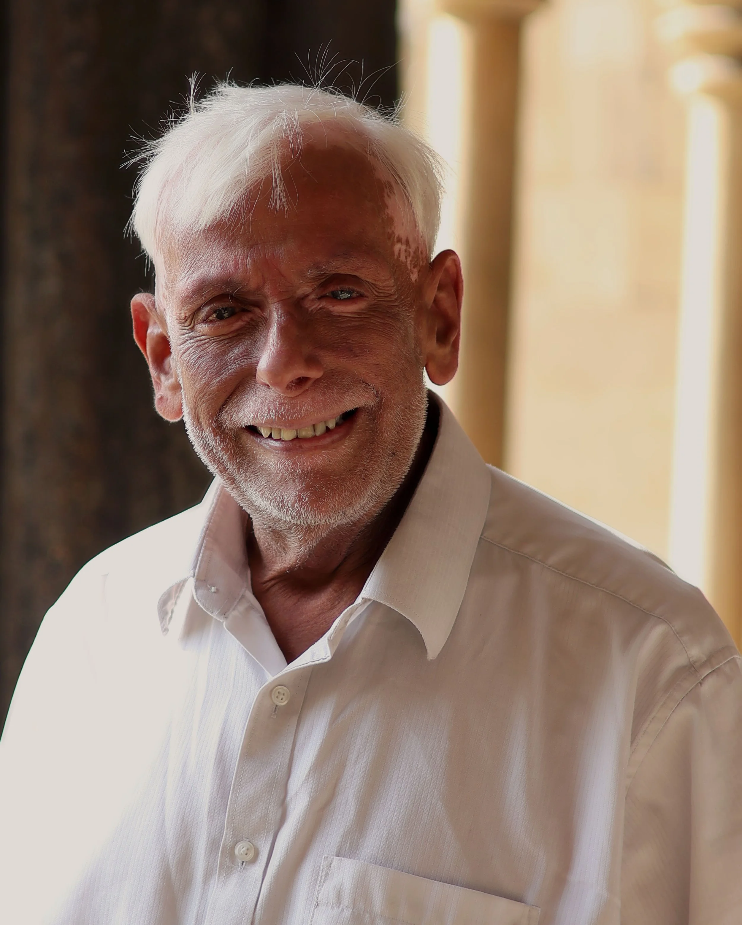 Close-up of an elderly man with white hair, smiling, wearing a white collared shirt, in a well-lit indoor setting.