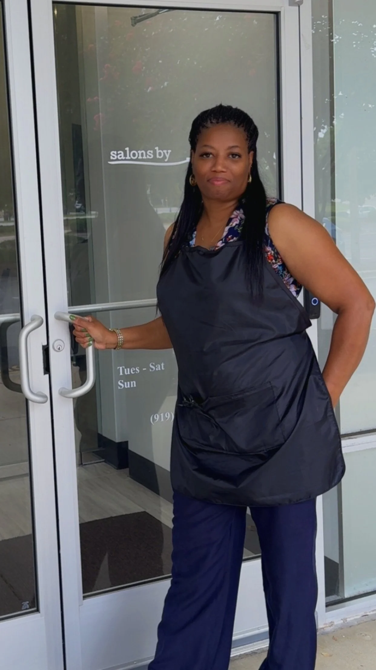 A woman with long, braided hair and wearing a navy blue apron opens a glass door to a salon, which is open Tuesday through Saturday, located in North Carolina.