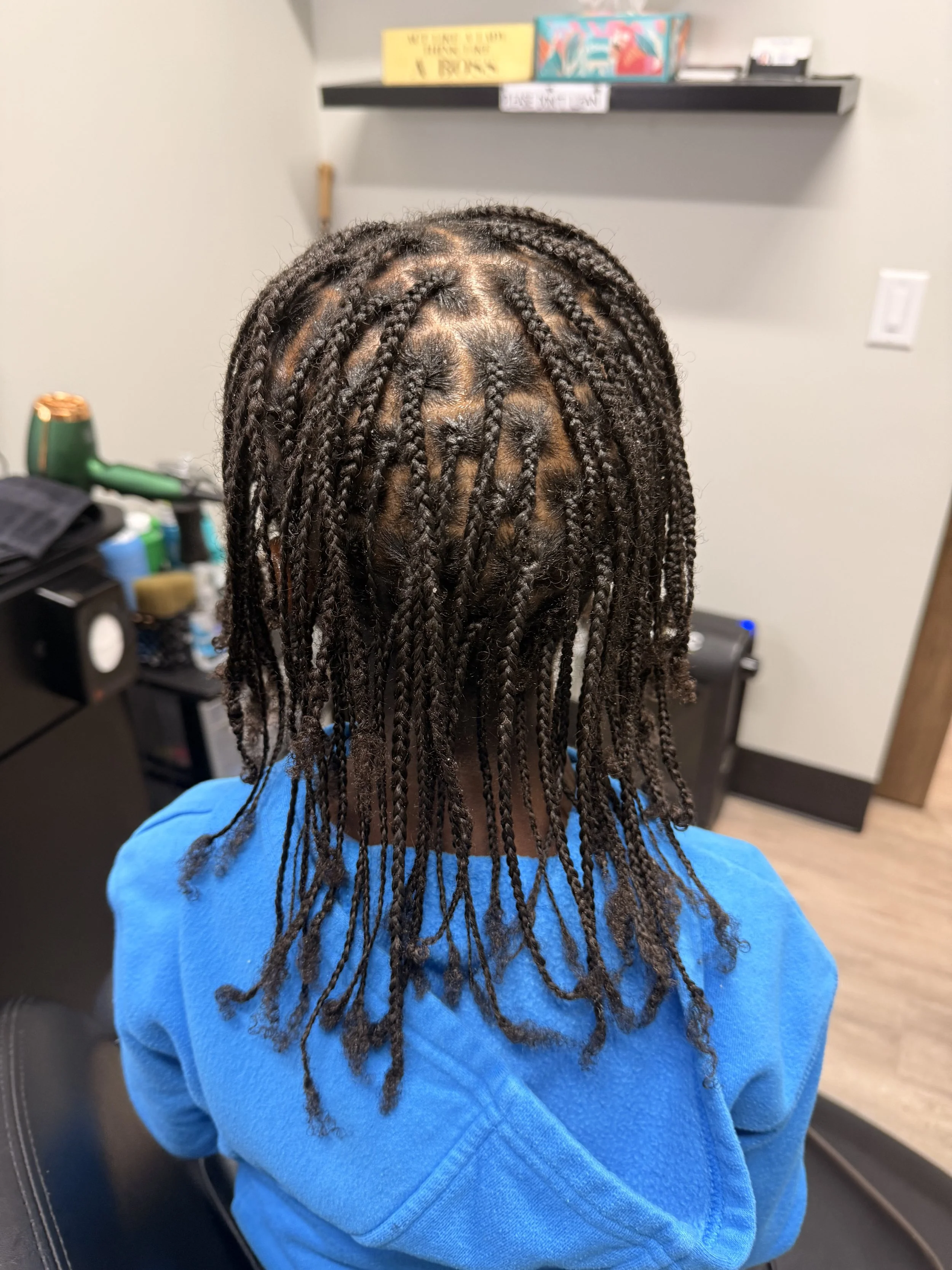 Back view of a person with freshly twisted locs hairstyle, wearing a blue shirt, sitting in a room with shelves and electronic equipment.