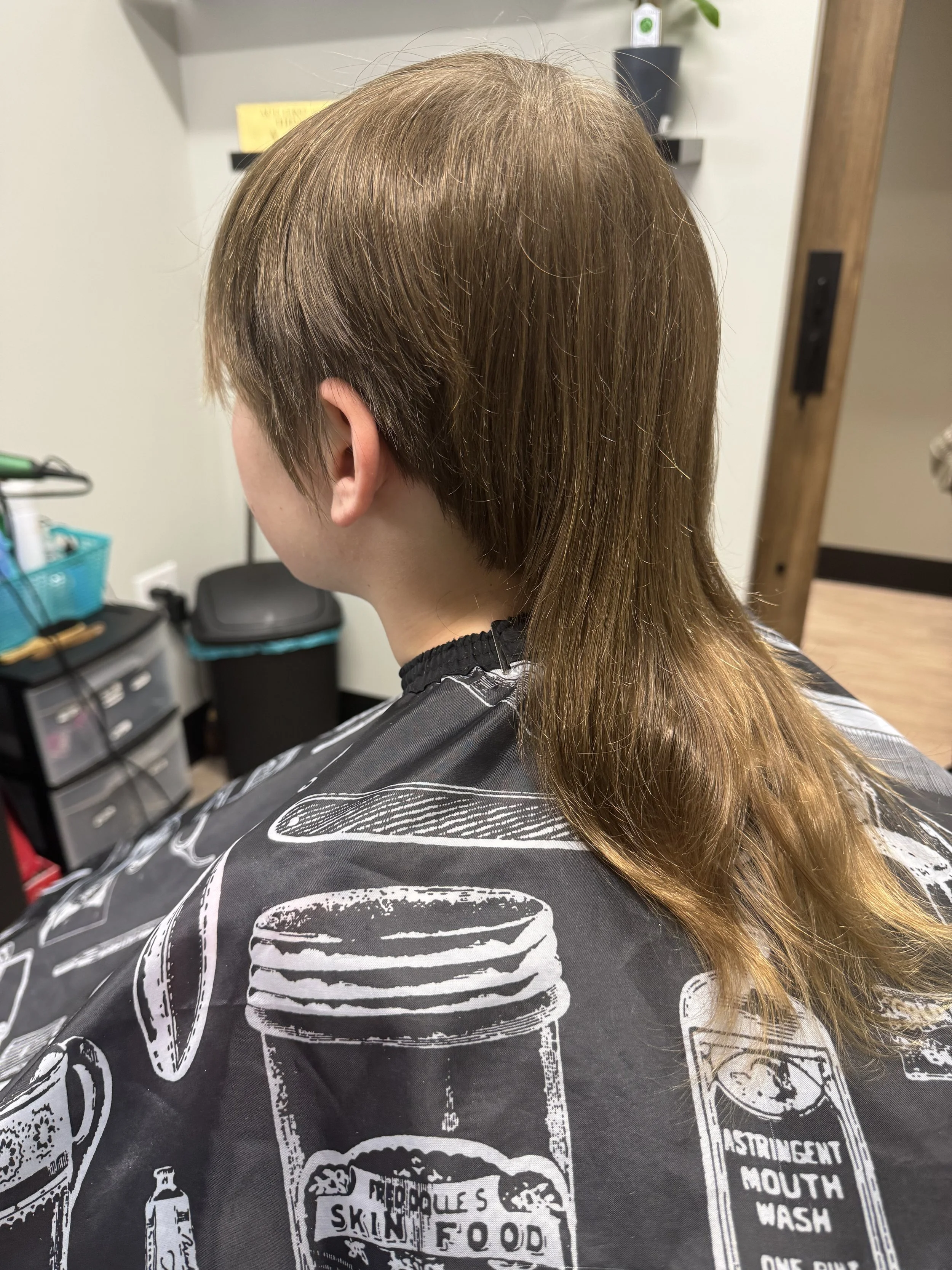 Back view of a person sitting in a salon chair with a brown, shoulder-length haircut, wearing a black salon cape with white illustrations of jars and bottles, including one labeled 'People's Skin Food' and another labeled 'Stringent Mouth Wash'.