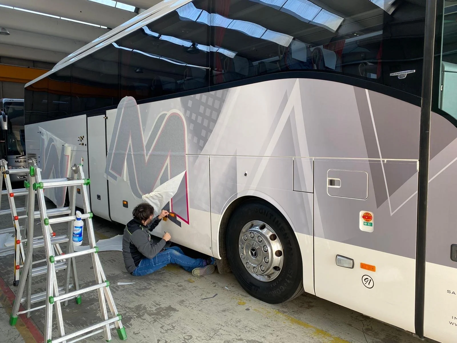 Una persona sta applicando una vernice o adesivo su un autobus bianco con parte di design geometrico. Sul lato dell'autobus ci sono diverse finestre e accessori, con una scala vicina.