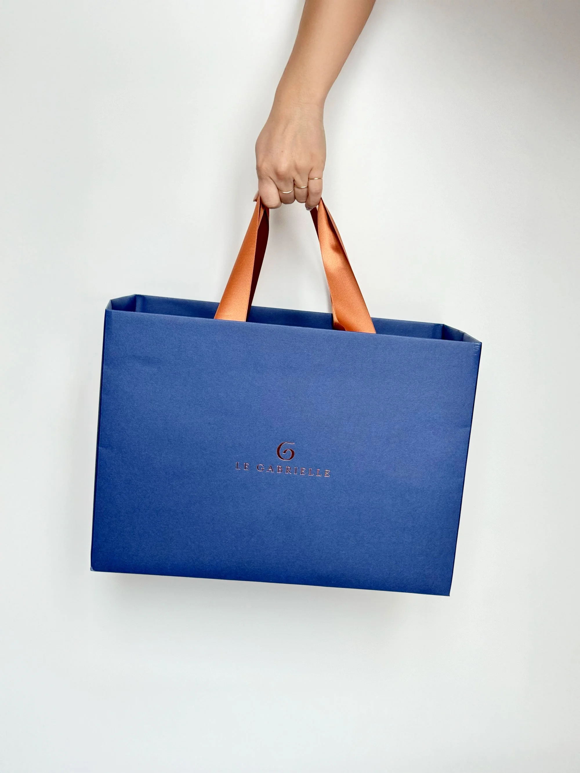 Mano che tiene una borsa blu con manici color arancio e il logo 'Le Gabrielle' al centro, contro sfondo bianco.