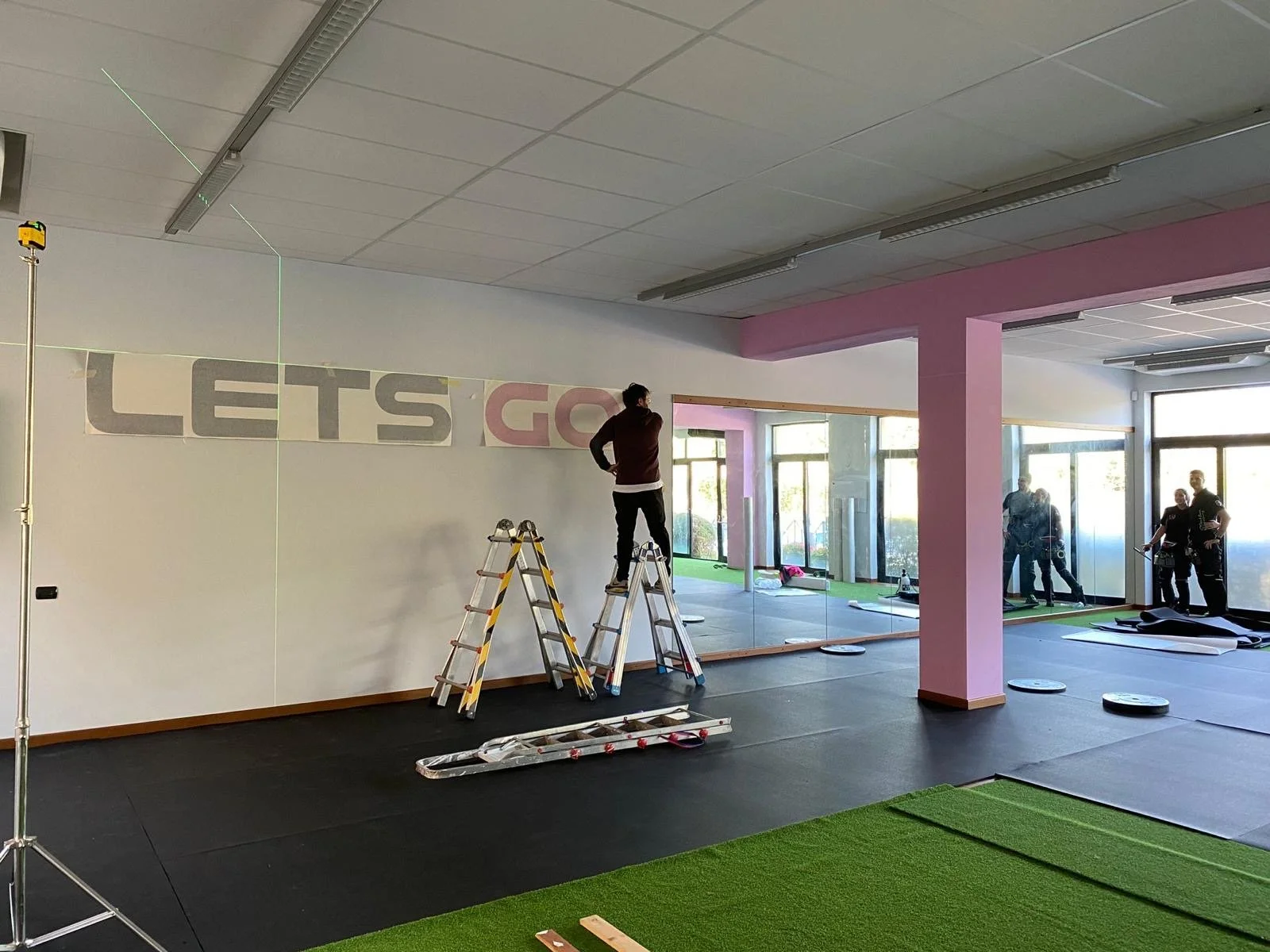 Interno di una palestra in fase di allestimento, con persone che sistemano un grande banner sul muro, attrezzi e pannelli in attesa di essere posizionati.