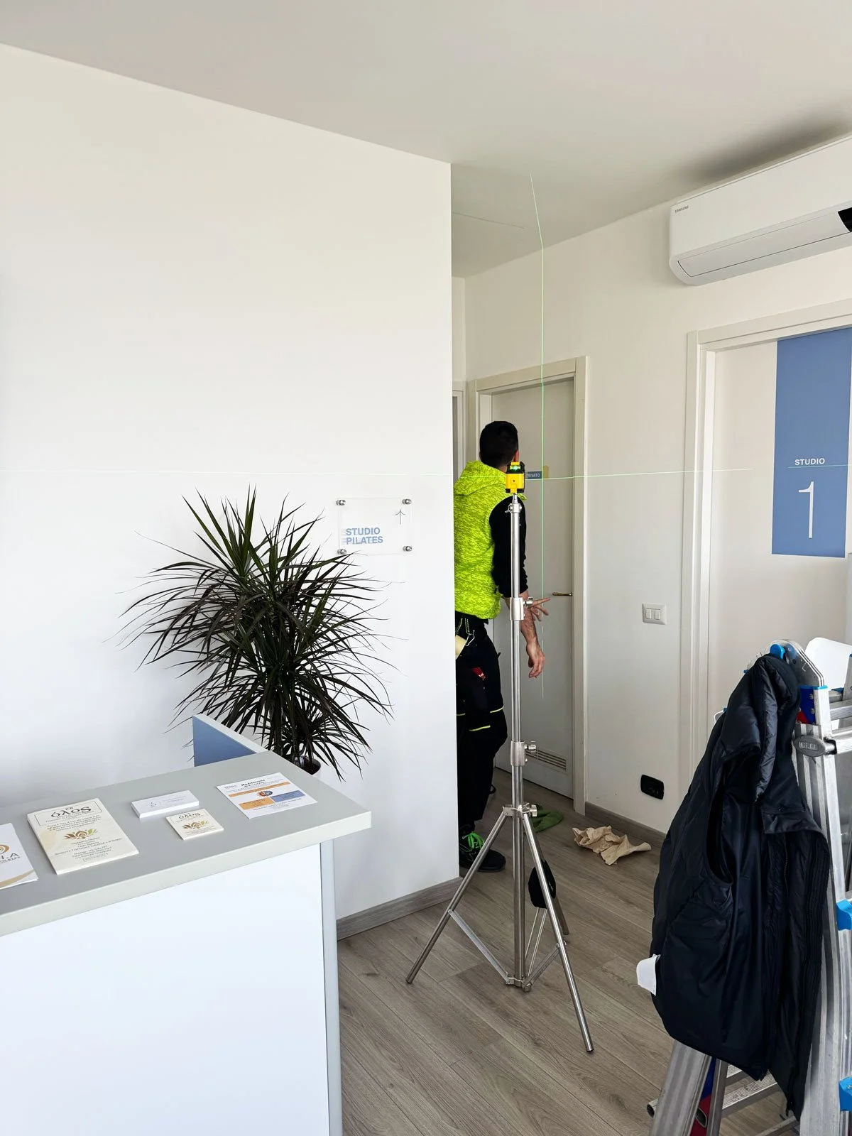 Interno di uno studio con un operatore che utilizza uno strumento di misurazione vicino alla porta. Tavolo con brochure e cartellino, piante, e altri attrezzi in vista.