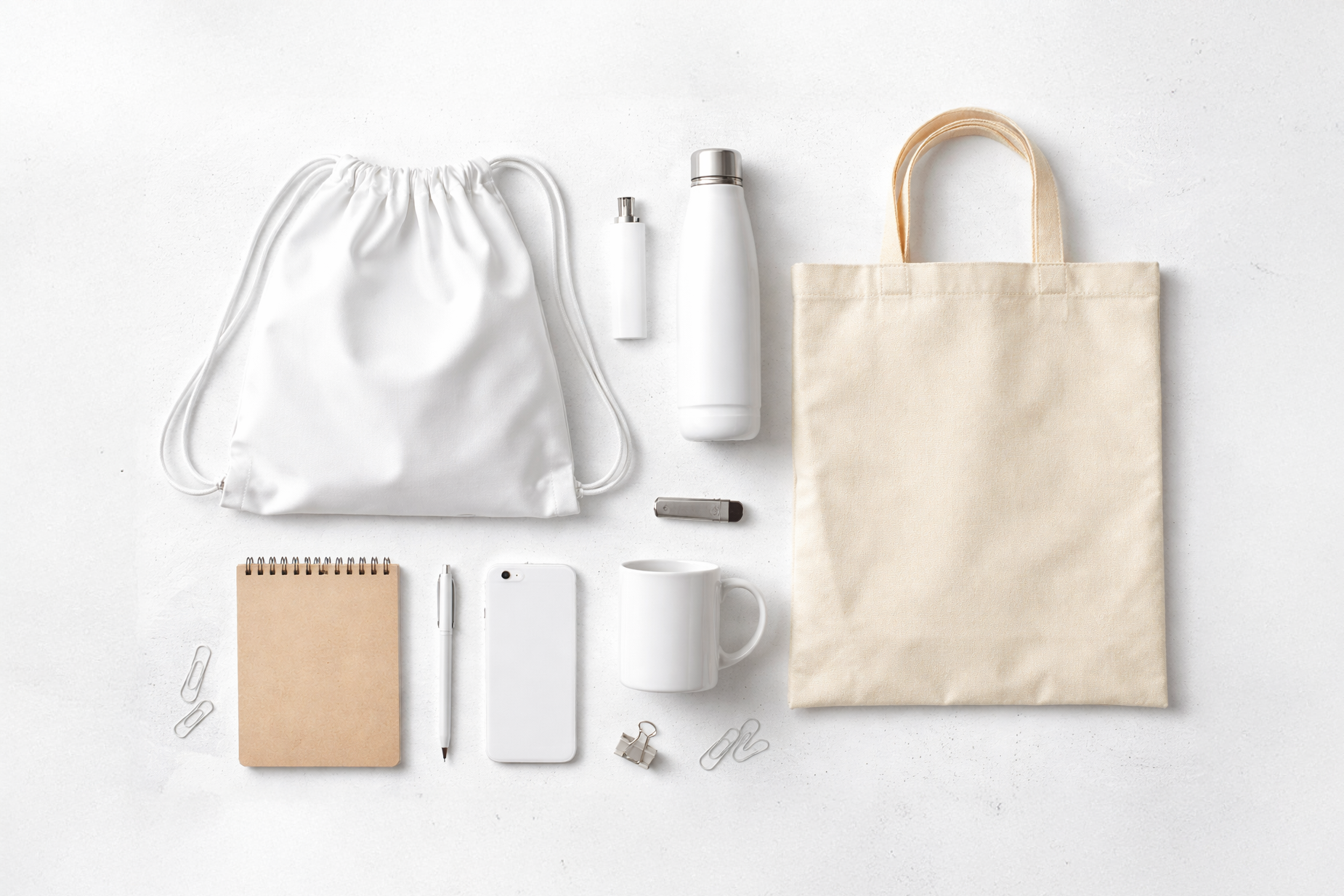 Gadget personalizzabili su una superficie bianca, inclusi una borsa in tessuto, una borraccia, una crema o spray, un tiralatte, un notebook, una penna, uno smartphone, una tazza, una clip, quattro graffette e alcuni fermagli.