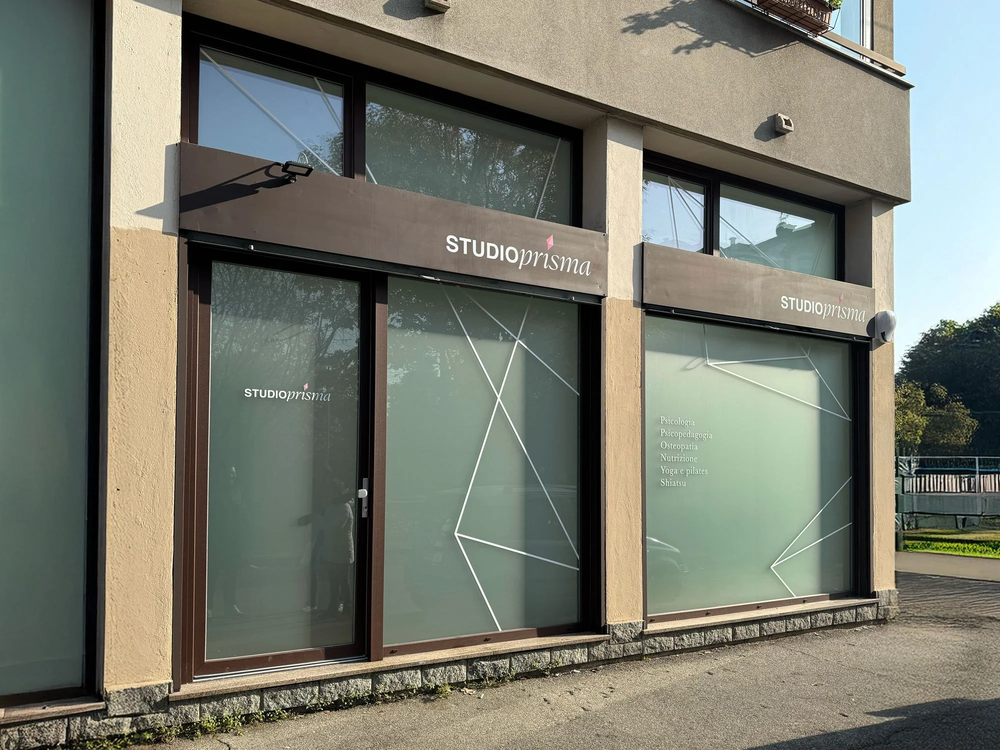Esterno di uno studio chiamato STUDIOprisma con vetrine sfumate e adesivi che indicano servizi come psicologia, psicopedagogia, osteopatia, nutrizione, yoga, pilates e shiatsu.