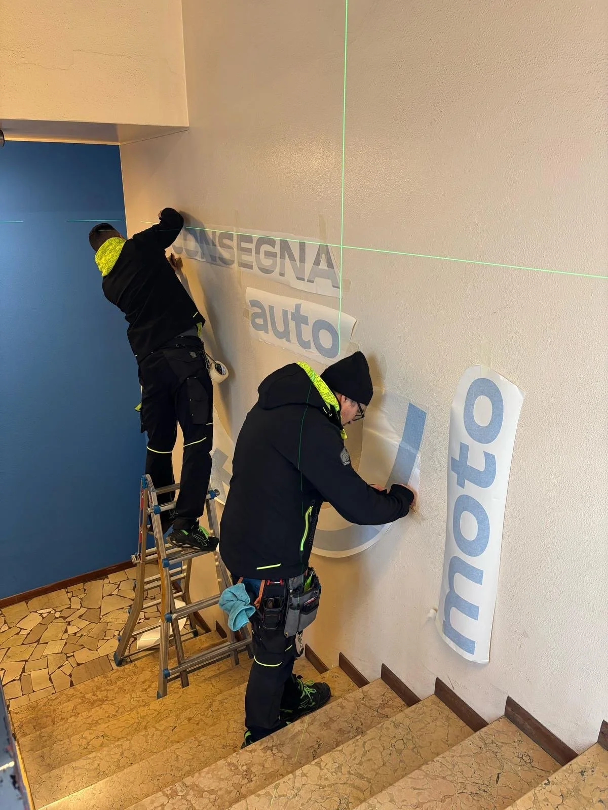 Due persone stanno attaccando cartelloni pubblicitari su una parete di un atrio. Una persona è sulla scala, l'altra sta in piedi a terra, entrambe sono vestite con abbigliamento nero con dettagli fluorescenti.