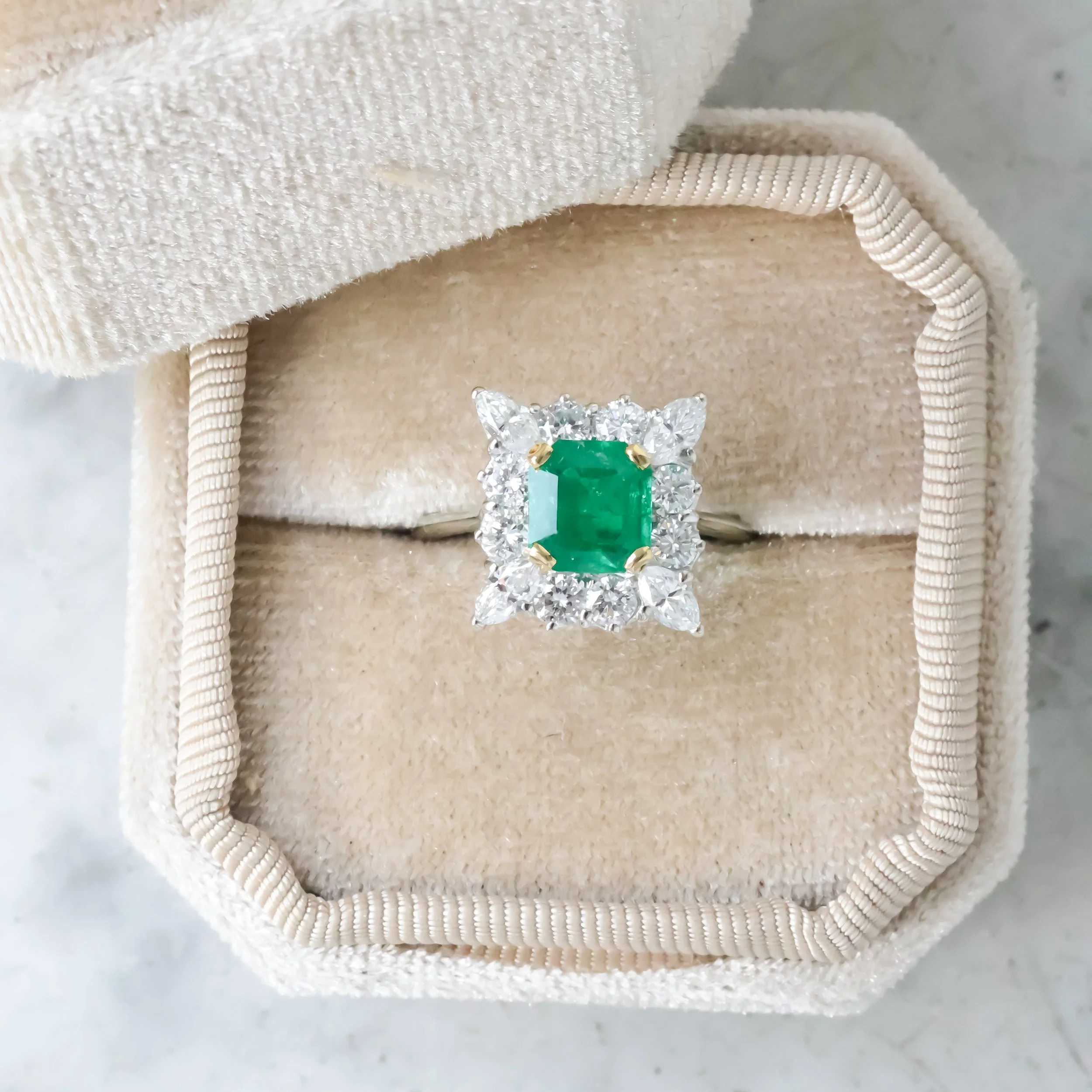 Bague avec une émeraude de forme carrée et entourée de diamants dans une boîte en velours beige.