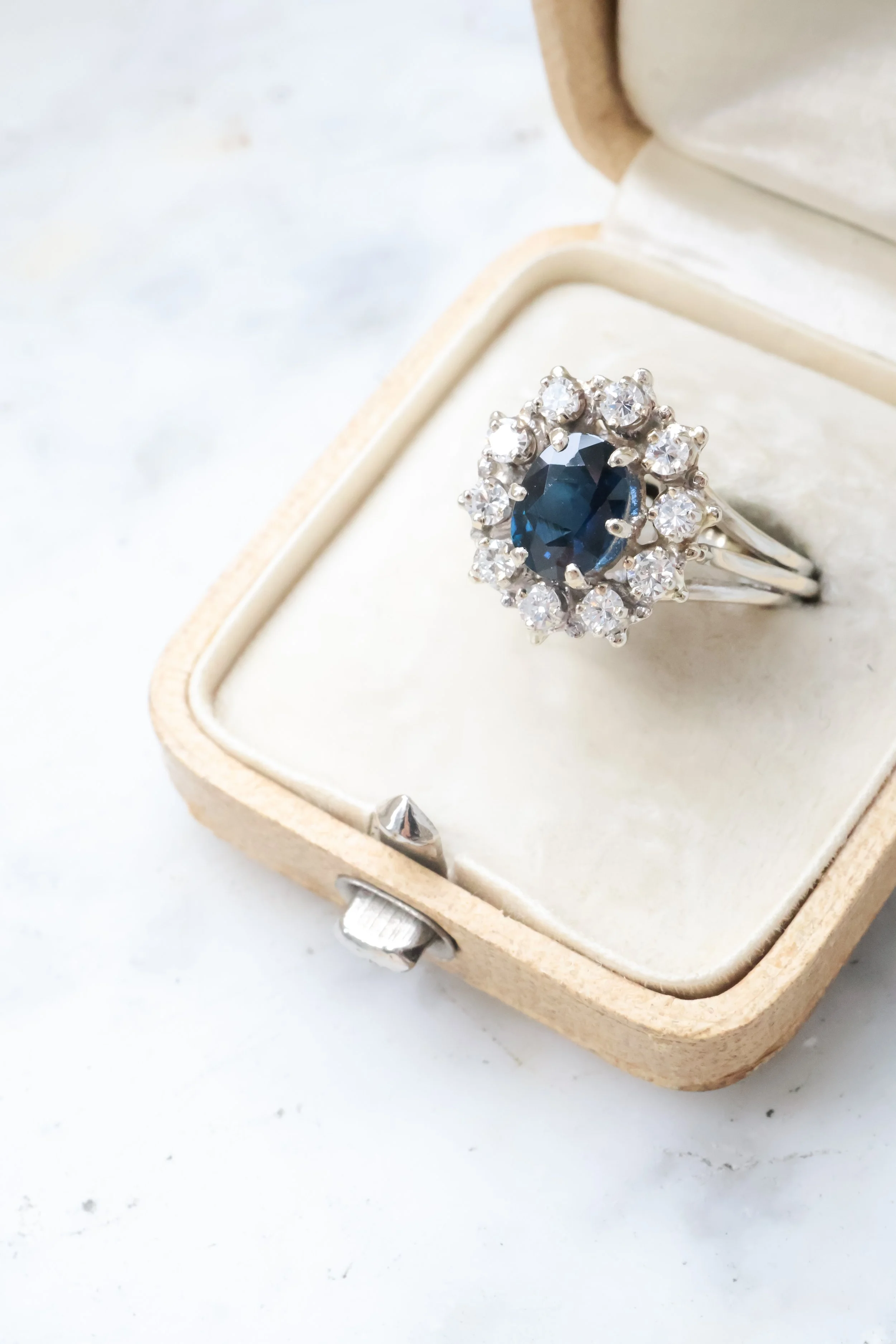 Une bague avec un gros saphir bleu entouré de diamants, posée dans un écrin en velours beige.