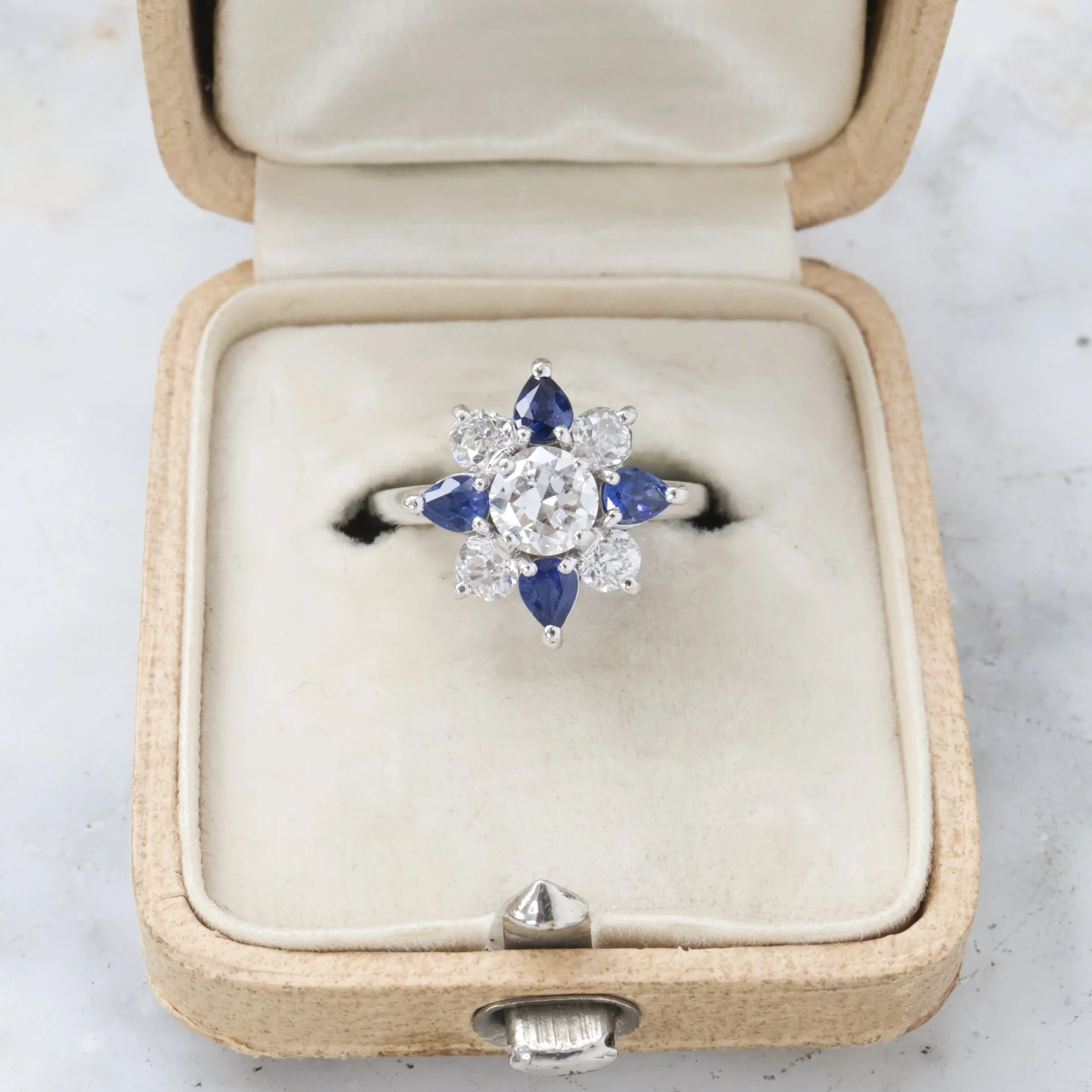 Bague en or blanc ornée de diamants et de saphirs bleus dans une boîte à bijoux vintage en velours blanc.