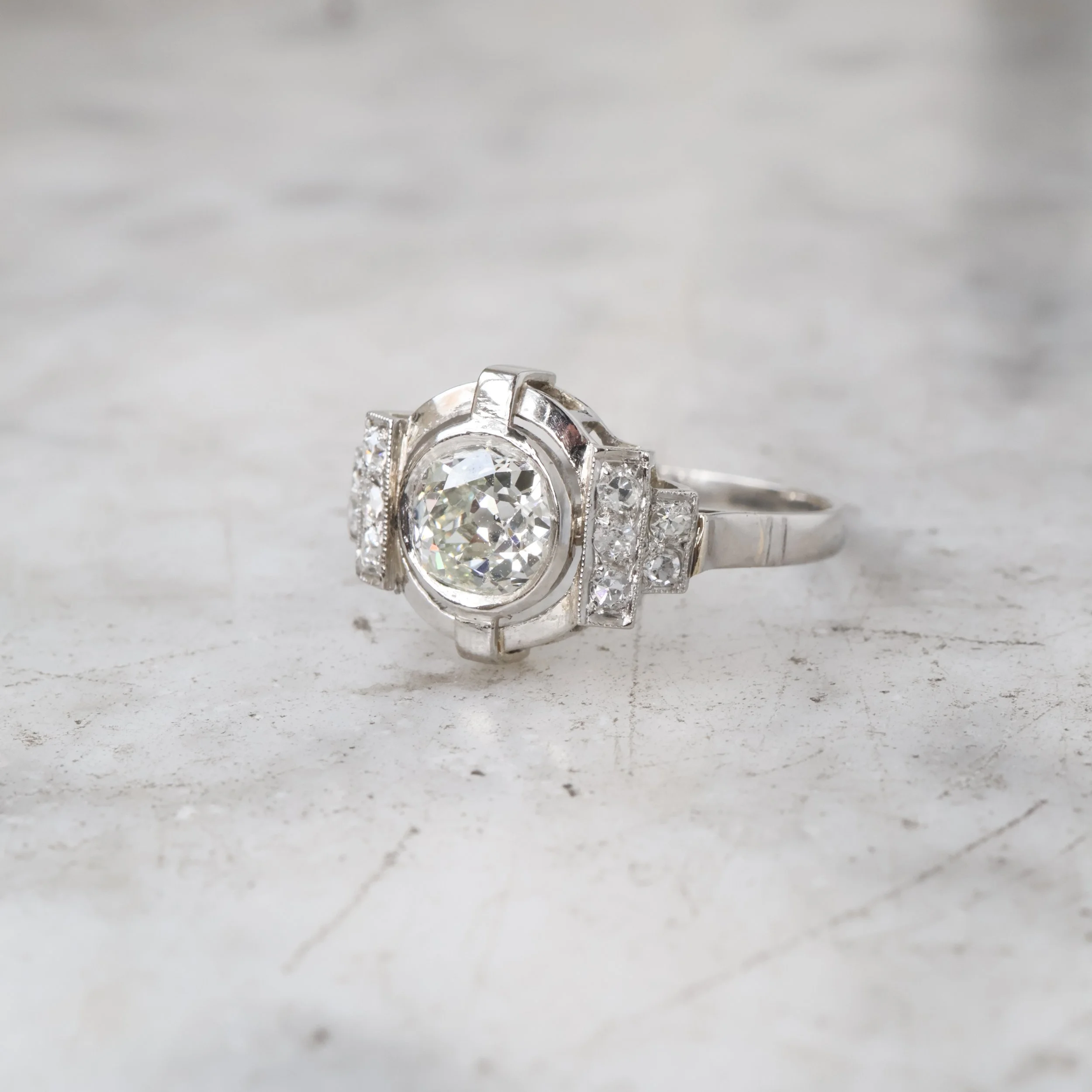 Bague en or blanc avec un grand diamant central entouré de petits diamants, sur un fond en surface grise