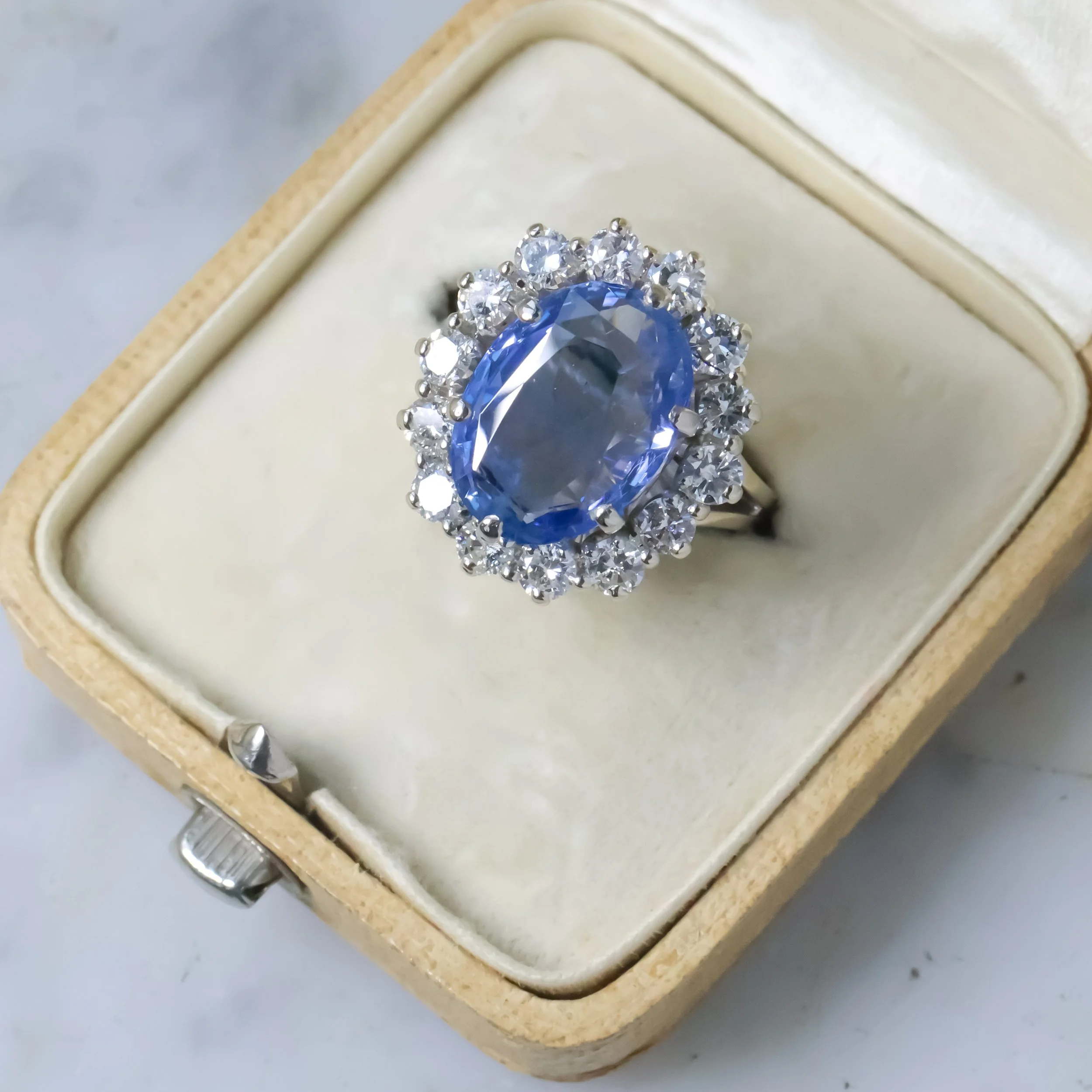 Bague avec une grande pierre ovale bleue entourée de petits diamants dans une boîte à bijoux beige.
