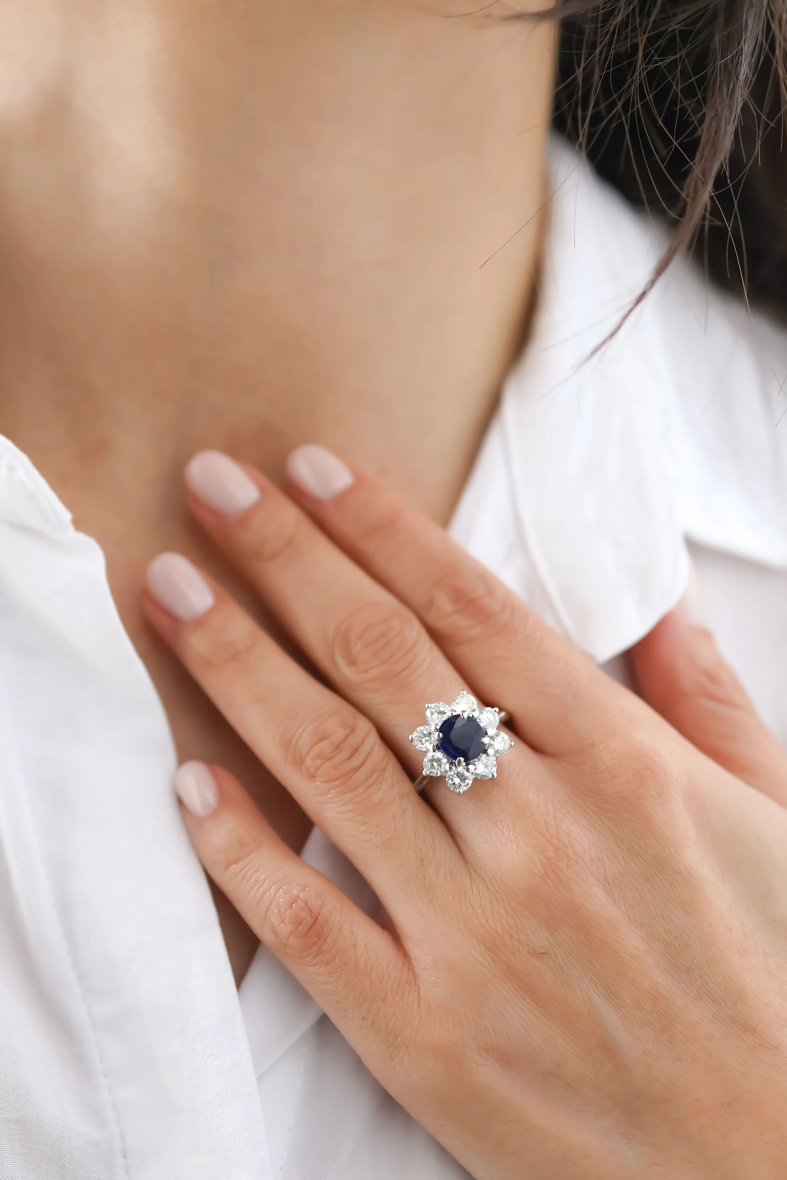 Une main portant une bague avec un grand saphir entouré de diamants, posée sur le poignet d'une autre personne, en arrière-plan une chemise blanche.