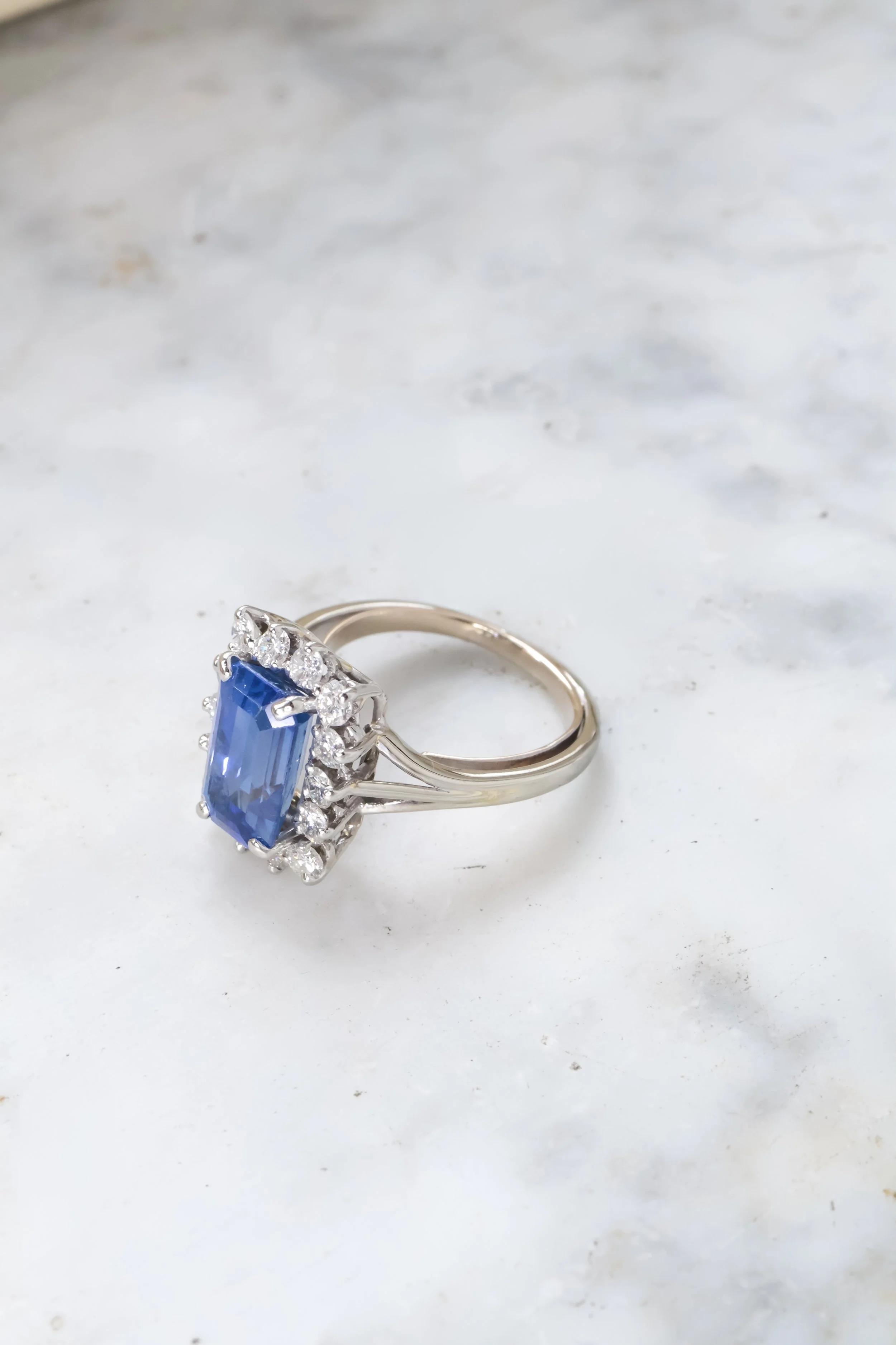 Une bague en or blanc avec un grand saphir bleu au centre entouré de petits diamants, posée sur une surface blanche