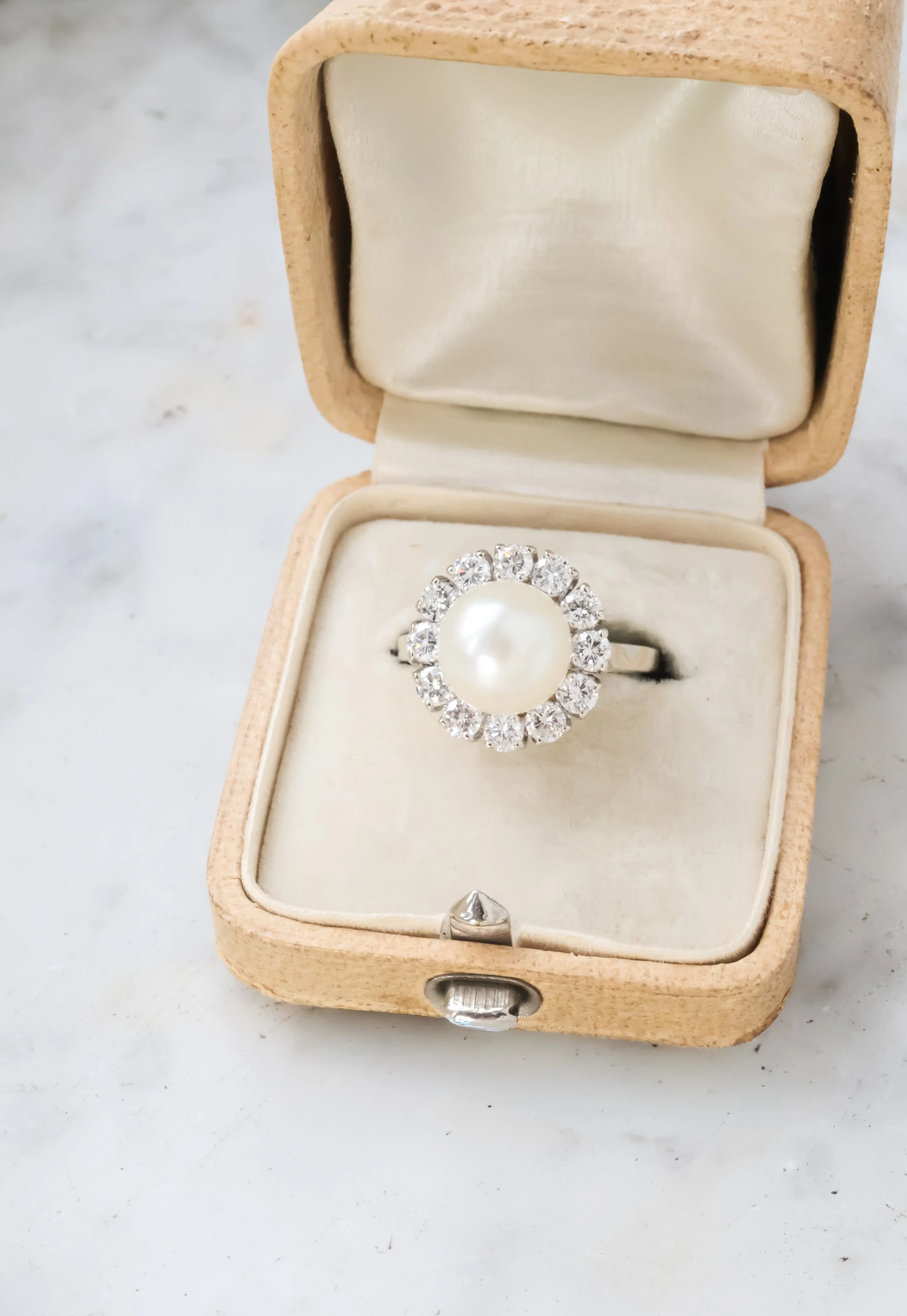 Bague ornée d'une perle centrale entourée de diamants, présentée dans une boîte en velours beige