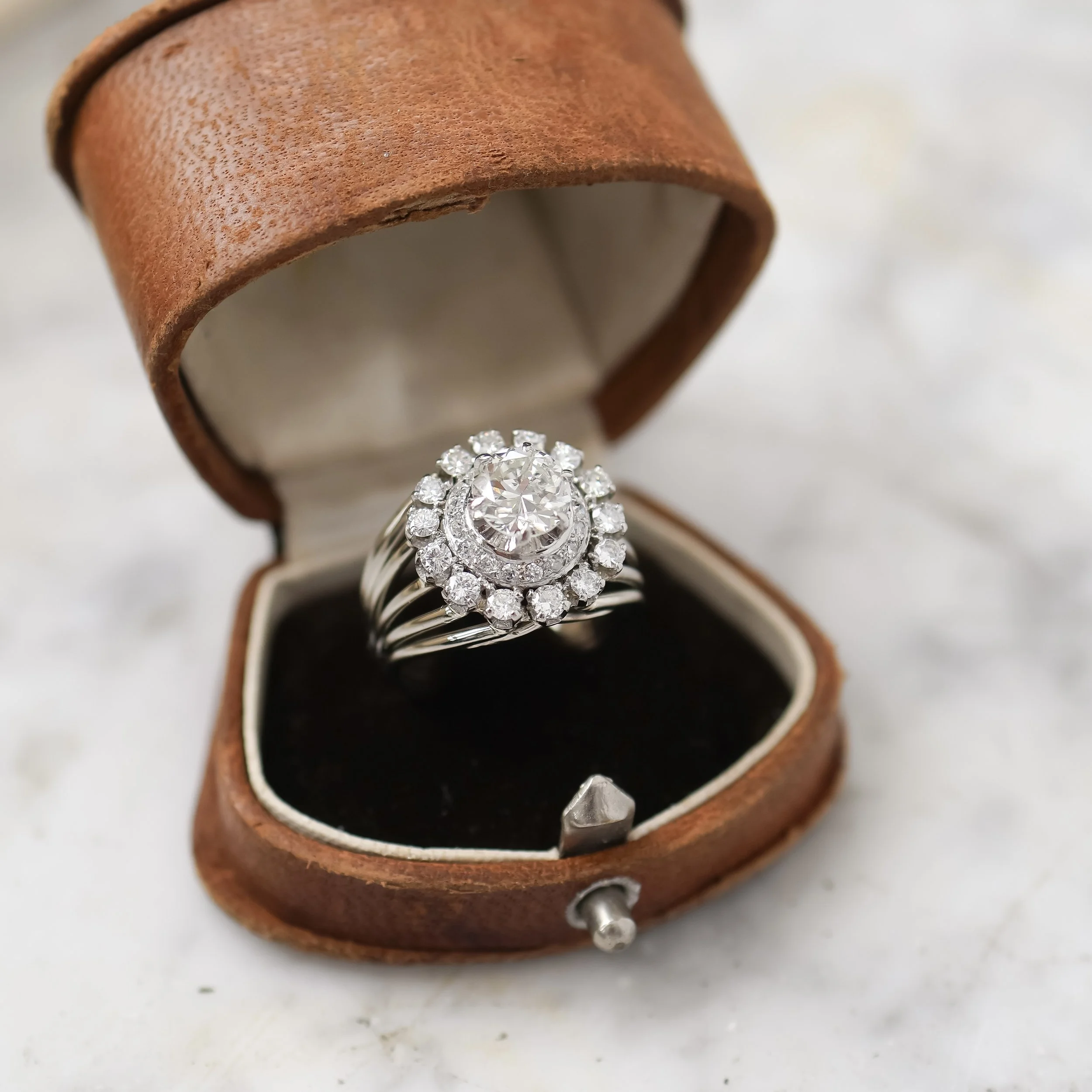 Bague en or blanc avec un grand diamant central entouré de petits diamants, dans une boîte à bijoux en cuir marron.