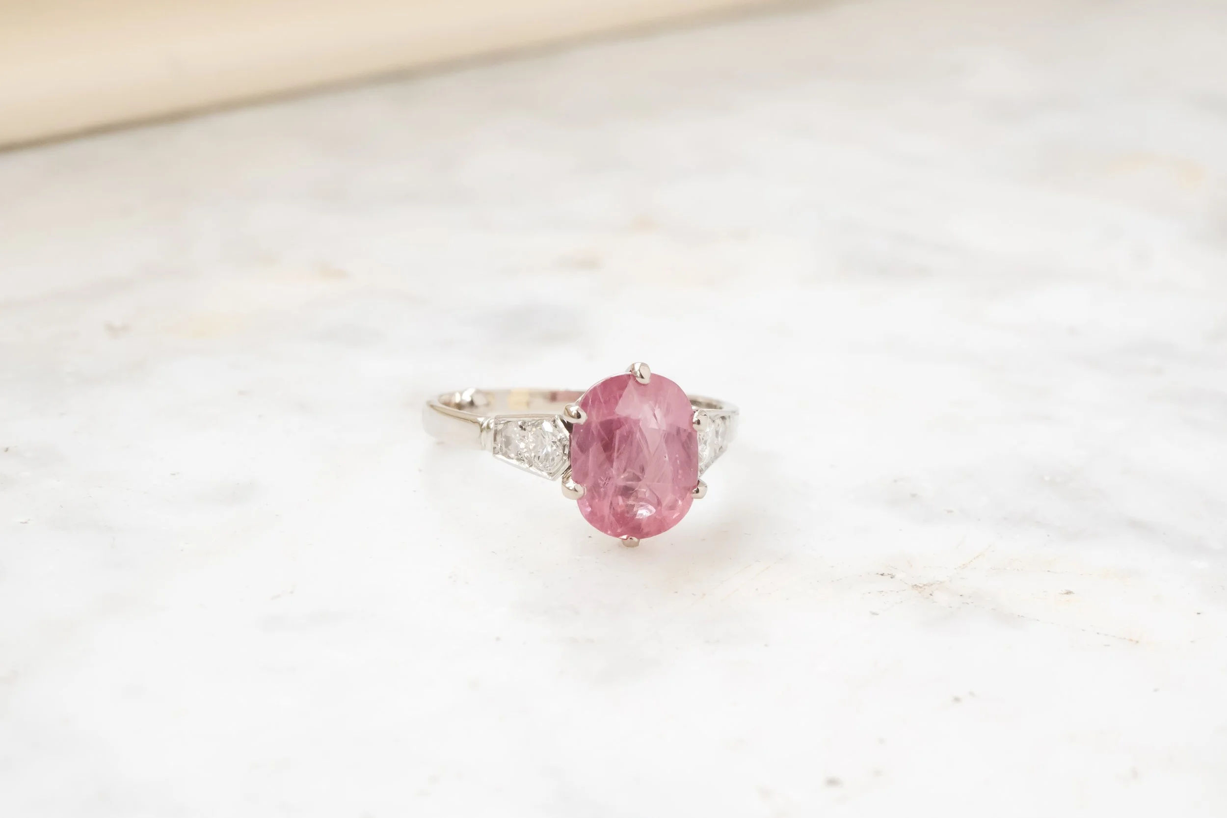 Bague avec une pierre centrale rose en forme d'ovale, entourée de deux petites pierres blanches de chaque côté, en métal argenté, posée sur une surface blanche.